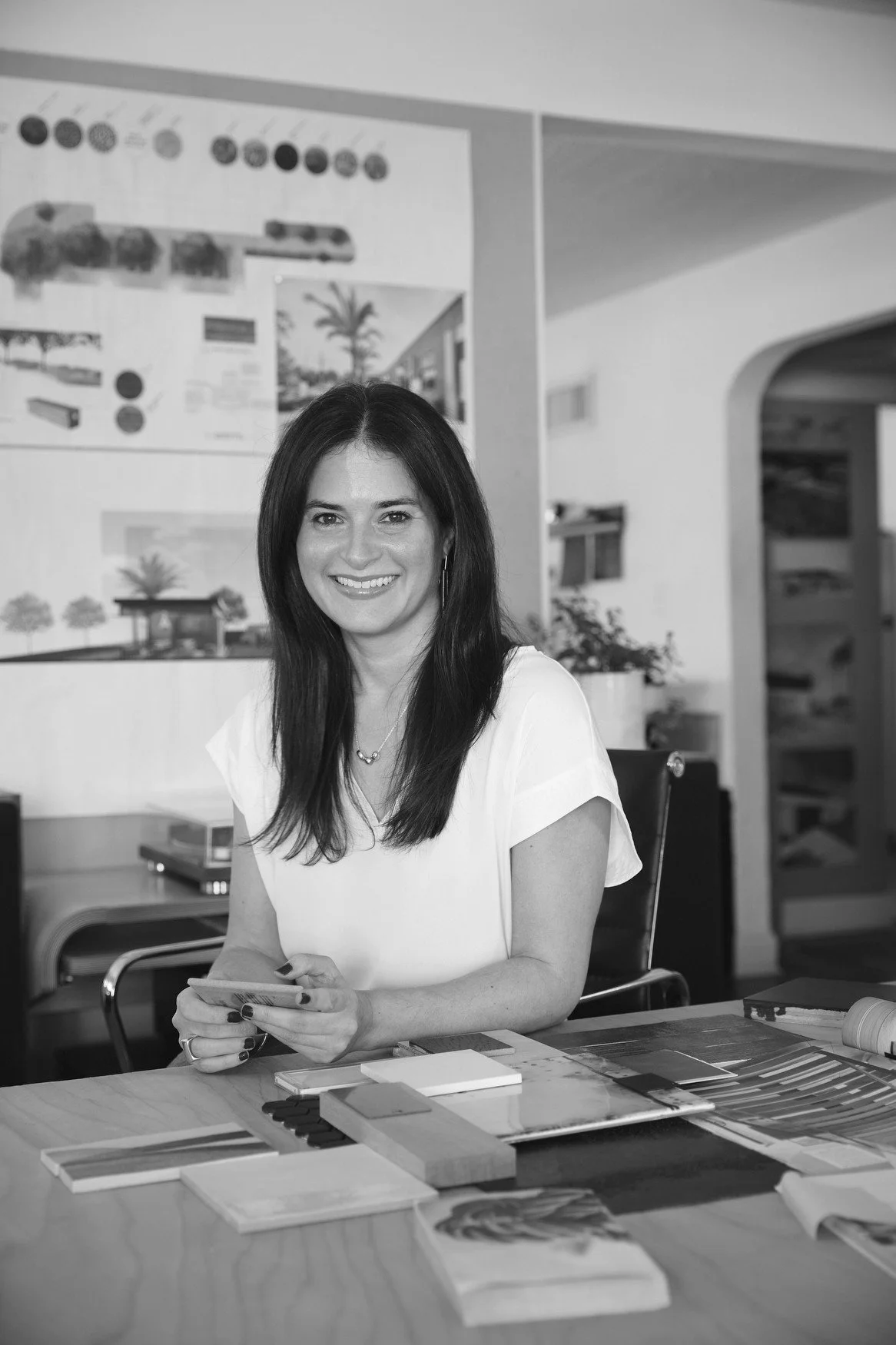 A woman with dark hair, wearing a white blouse, sitting at a table with design and color samples, smiling and holding a piece of paper, in an office or design studio setting.