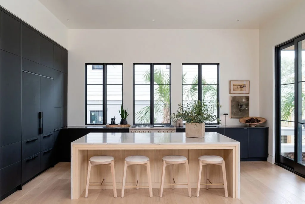 Modern kitchen with a white island, three stools, black cabinetry, and large windows showing palm trees outside.