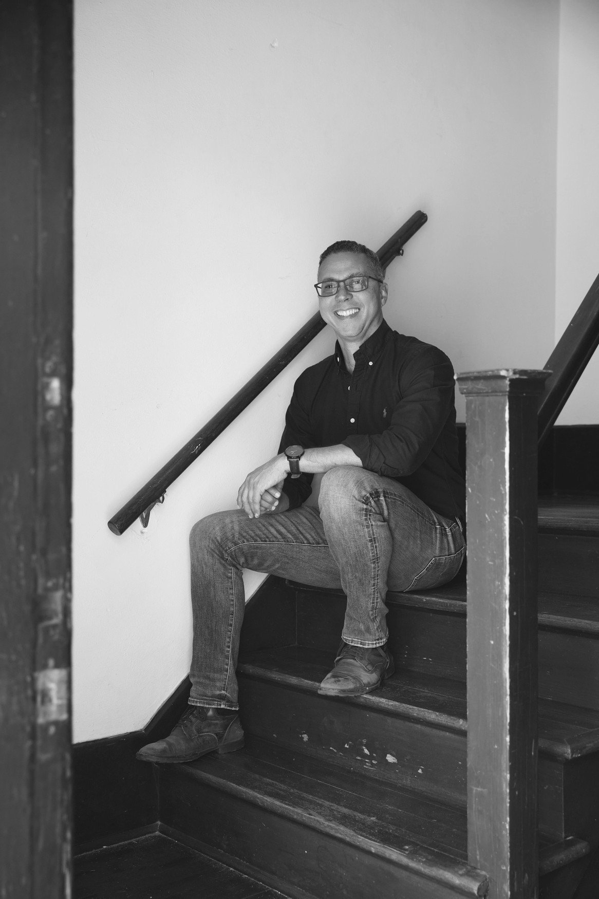 A smiling man wearing glasses, a black button-up shirt, and jeans, sitting on a staircase with a railing, indoors, in black and white.