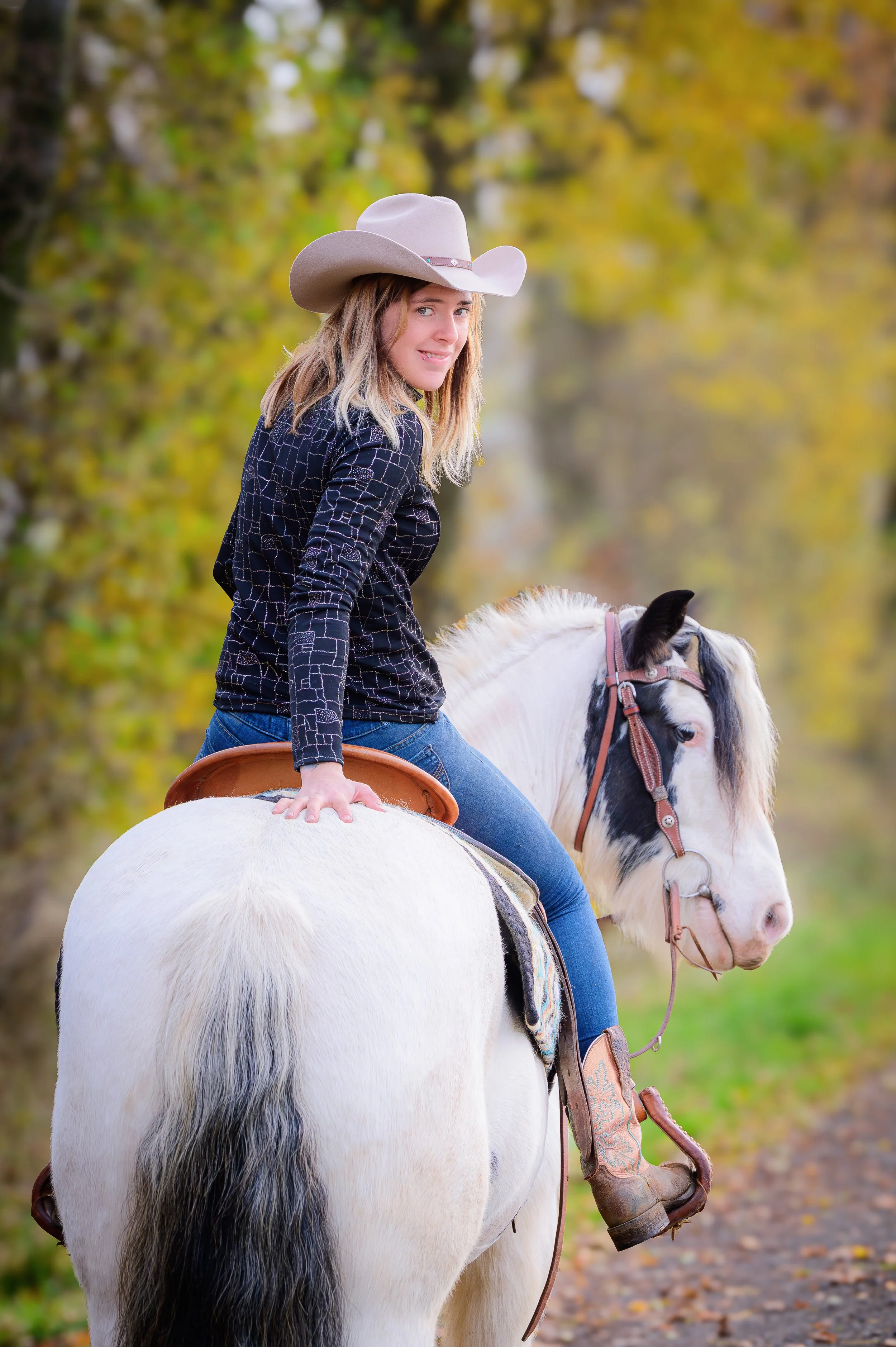 Eine junge Frau mit Cowboystiefeln, Jeans, einer schwarzen Jacke und einem Cowboyhut sitzt auf einem weißen Pferd mit schwarzen Flecken, im Hintergrund sind Bäume mit gelbem Laub, vermutlich im Herbst.