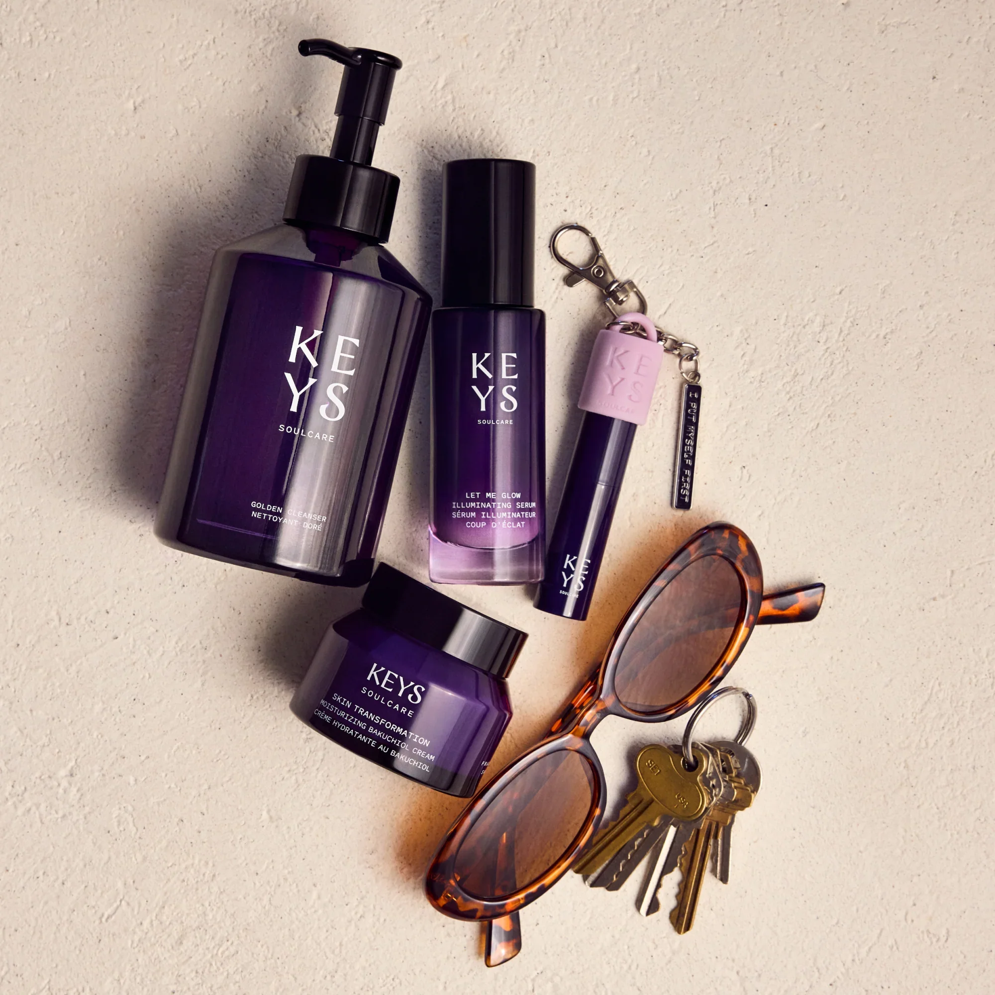 Collection of Keys Soul Carevskincare products including two purple bottles, a pink keychain with the word 'KEYS', and a pair of tortoiseshell sunglasses placed on a light-colored surface.