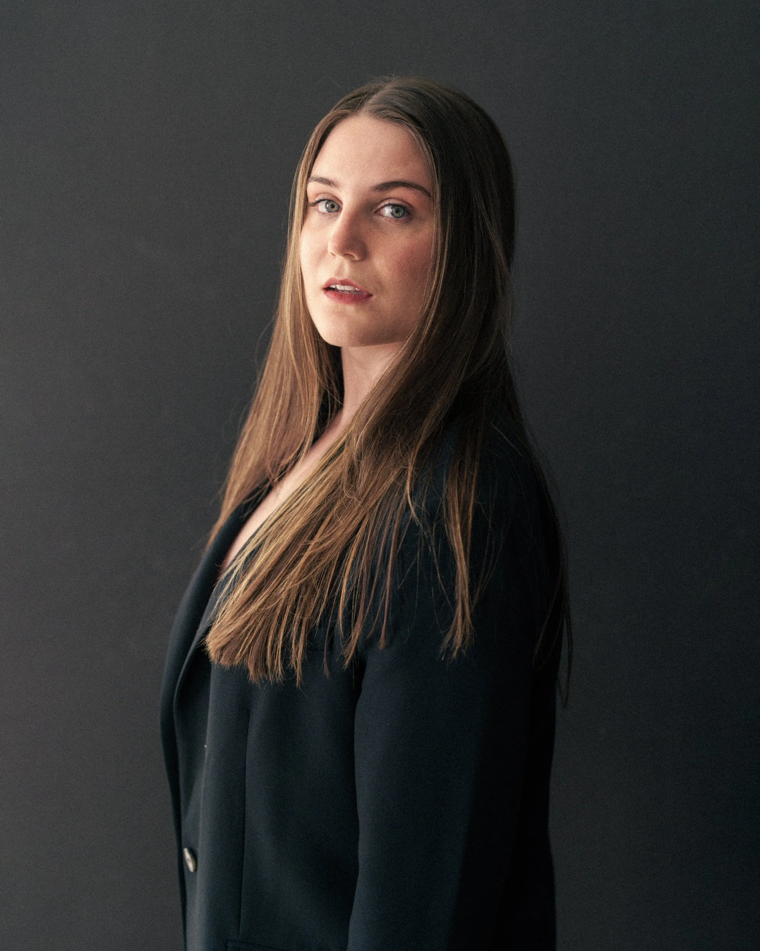 Portrait of a Marissa with long brown hair wearing a black blazer against a dark background.