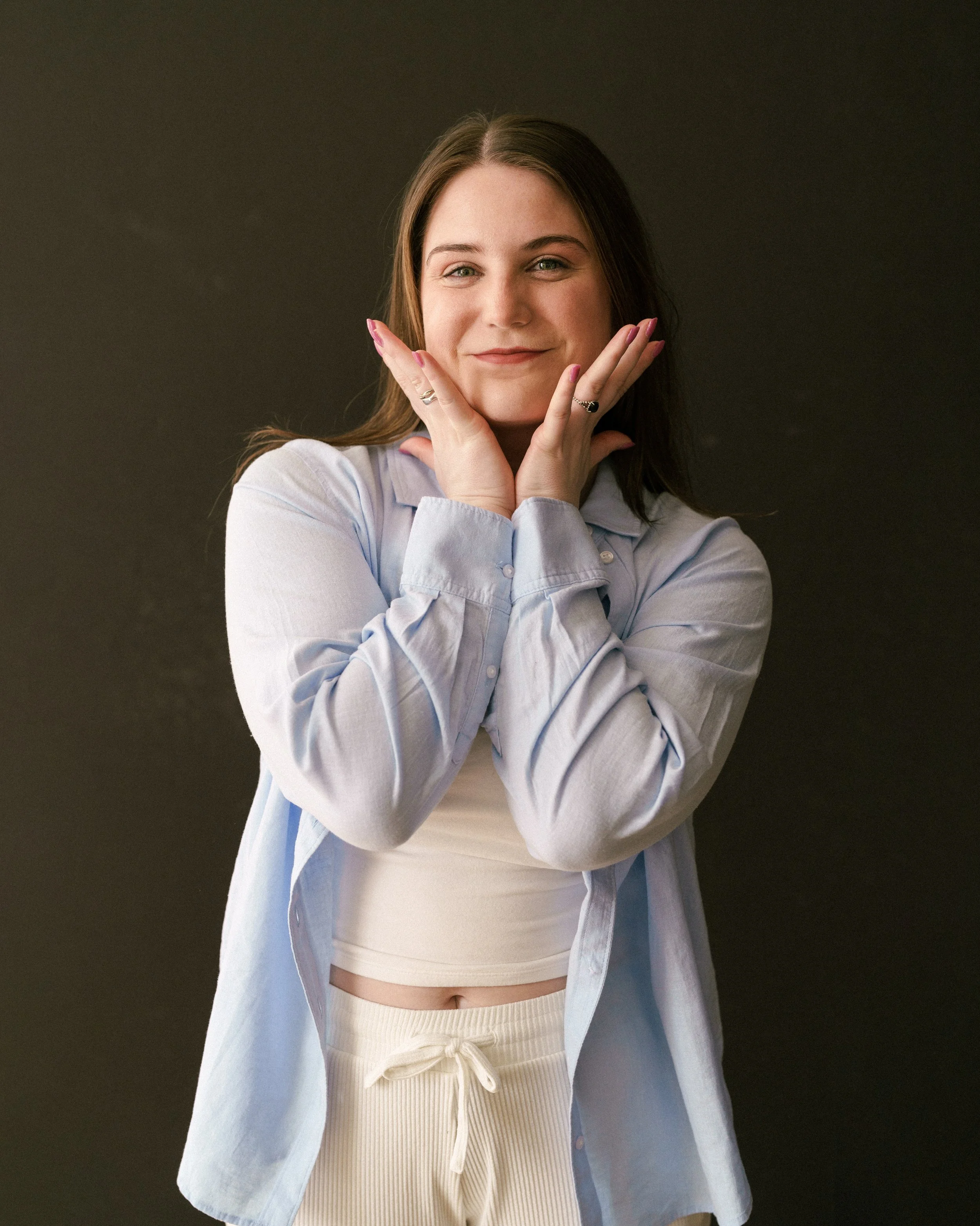 Marissa with long brown hair, smiling and touching her cheeks with her hands, wearing a light blue shirt, a beige crop top, and beige pants against a dark background.