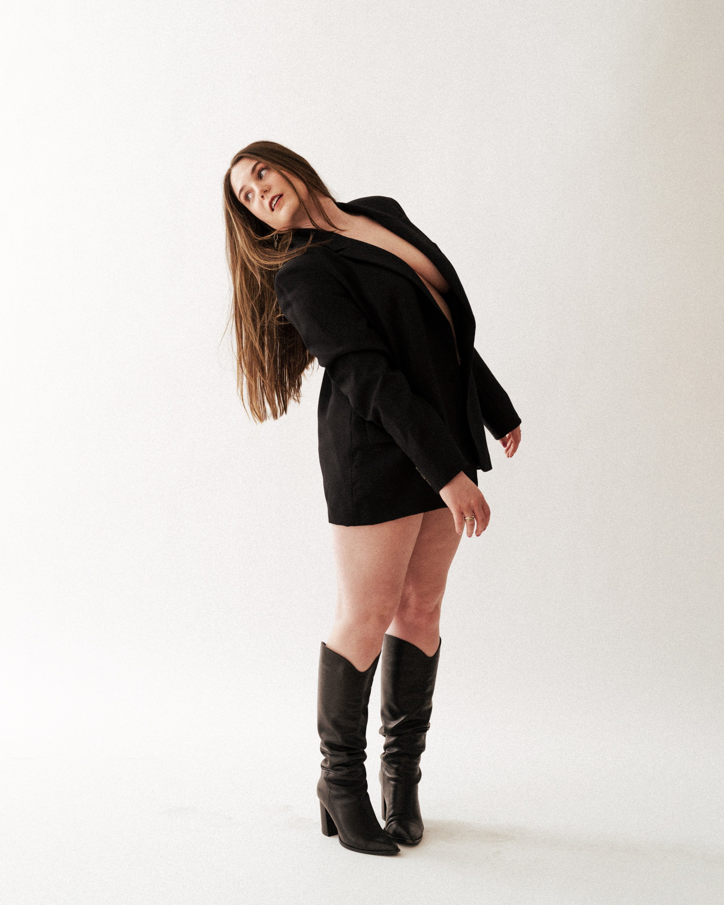 Marissa with long brown hair wearing a black blazer, shorts, and high-heeled boots standing against a plain background.