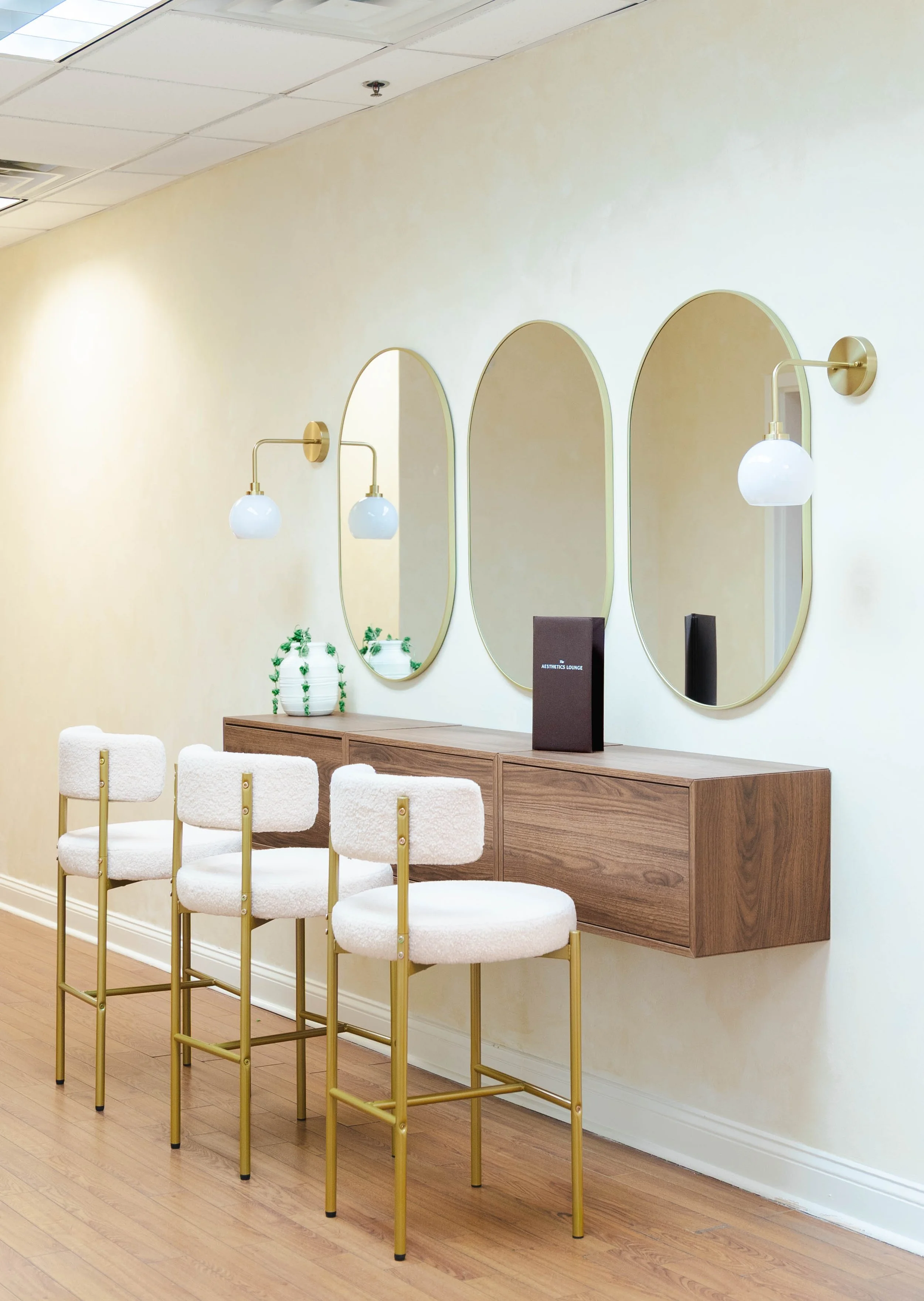 Interior of a modern salon or lounge area with a wooden counter, three white chairs with gold legs, three oval mirrors mounted on a beige wall, and two wall-mounted lamps with white globes.