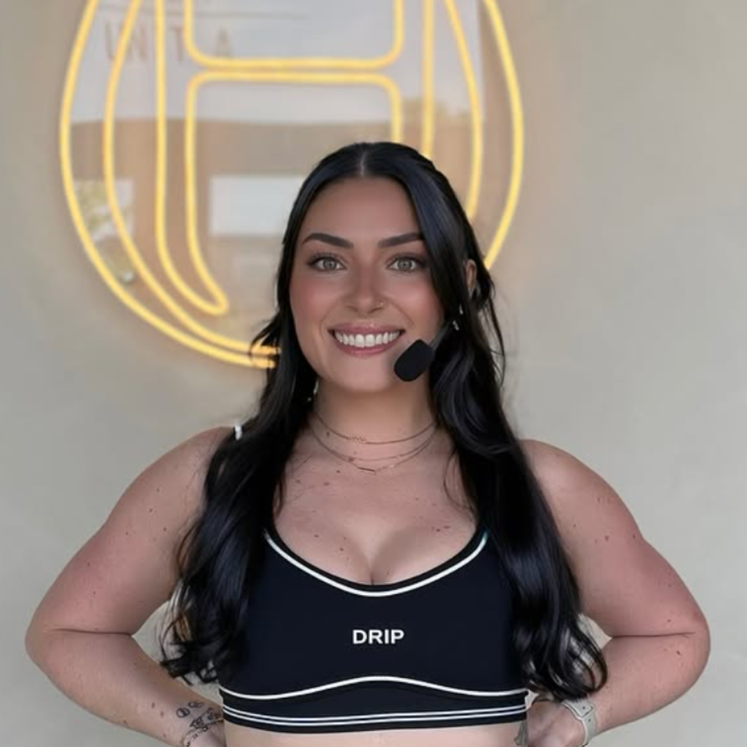 Girl with long dark hair wearing a black sports bra with white trim, smiling, standing in front of a neon banana sign, wearing a black headset microphone.