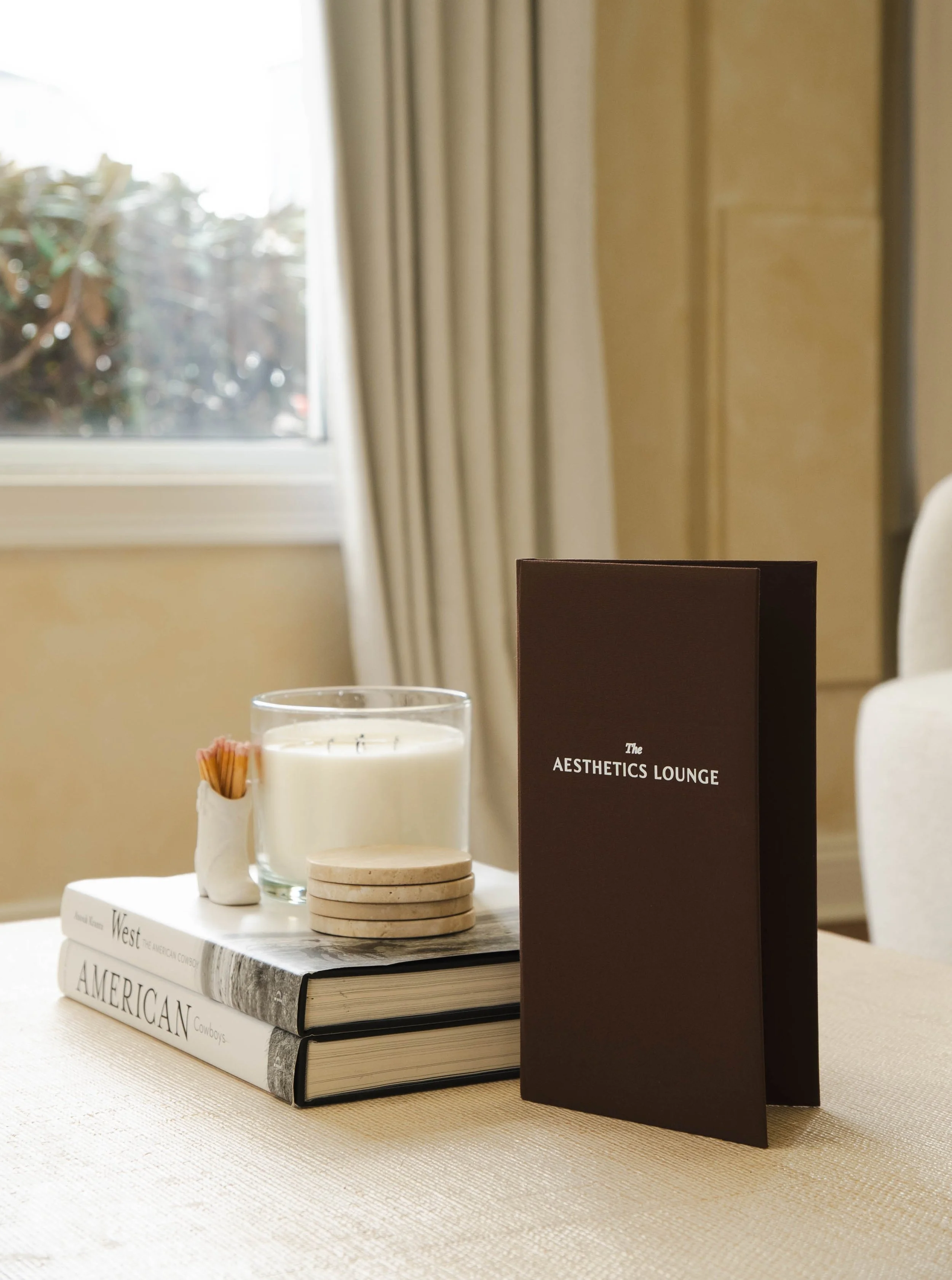A table setting with a glass candle, coasters, a menu titled 'The Aesthetics Lounge', and books about America, West, and Cowboys on a white tablecloth near a window with curtains.