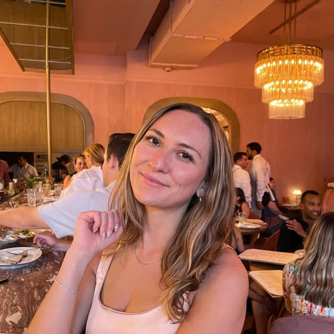 A woman with long wavy hair smiling at a restaurant, with people dining around her and warm lighting fixtures hanging from the ceiling.