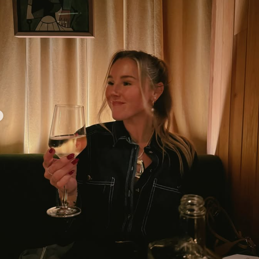 A woman sitting in a cozy, dimly lit room holding a glass of white wine with a satisfied expression.