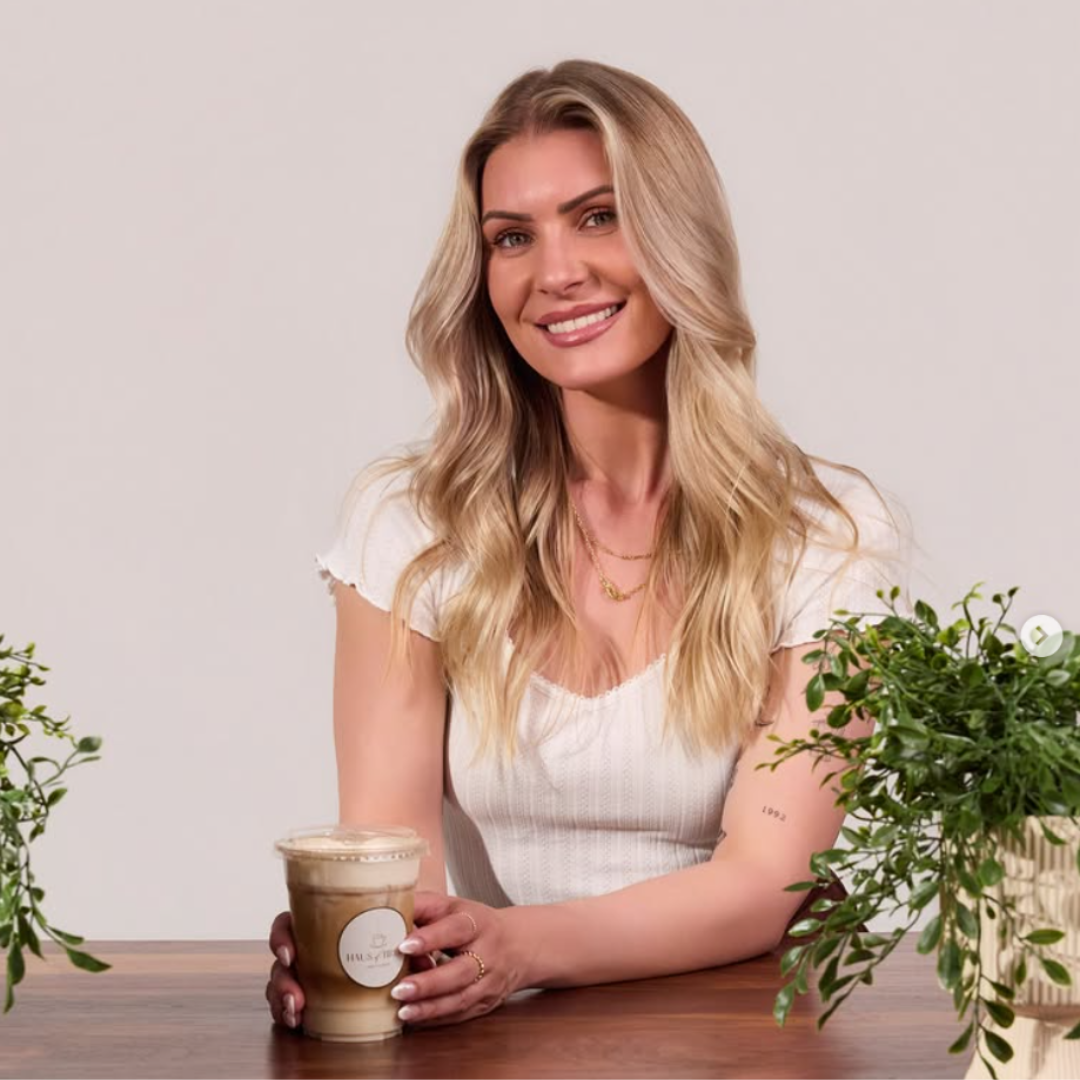 A blonde woman smiling, holding a coffee drink, sitting at a wooden table with green plants.