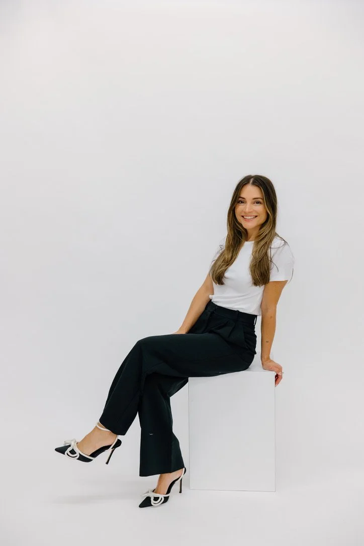 A woman with long brown hair smiling, sitting on a white box in front of a plain white background. She wears a white short-sleeve top, black high-waisted pants, and black-and-white high-heeled shoes.