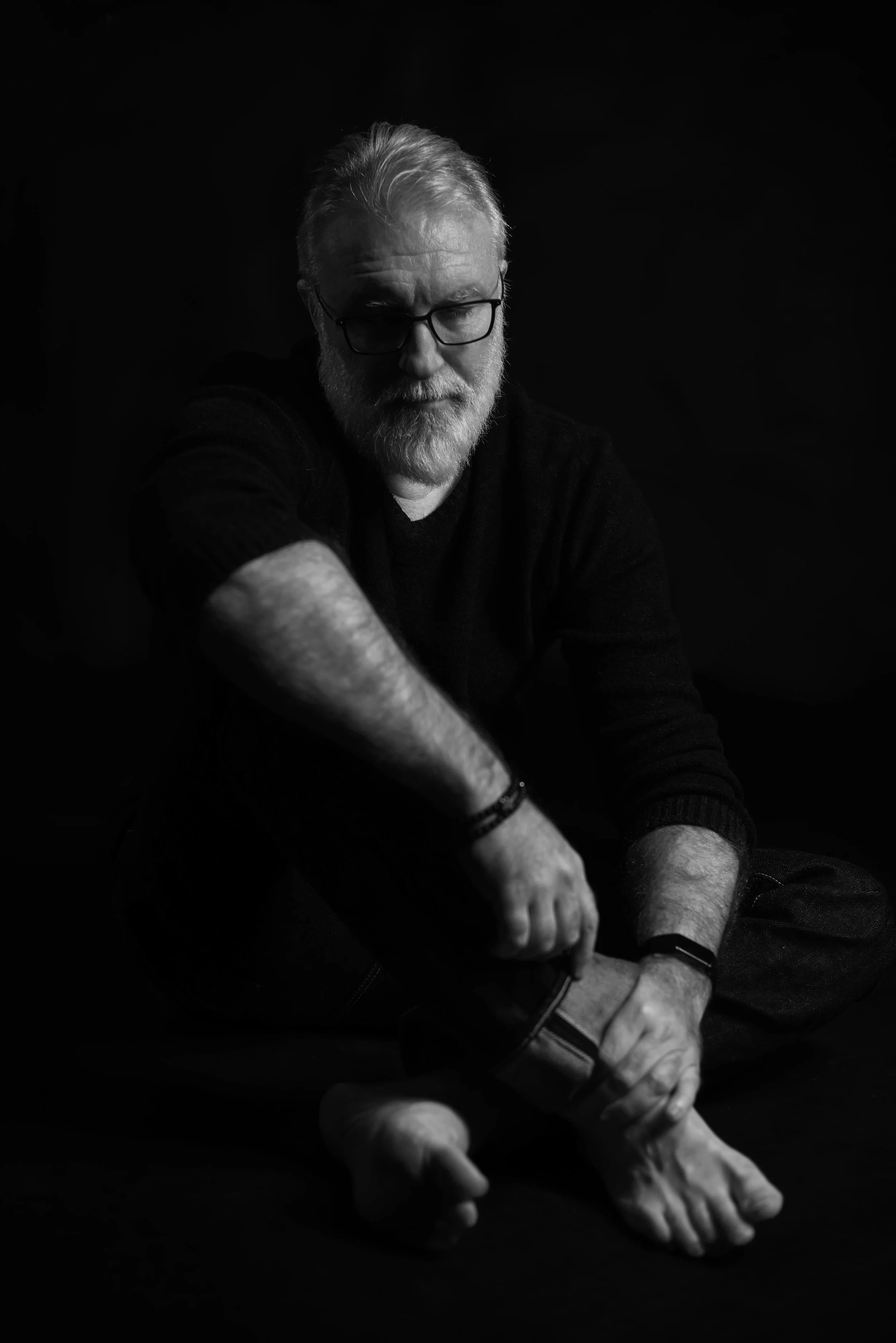 Uomo anziano con barba e occhiali neri seduto a terra su sfondo nero, che si piega in avanti con un braccio al ginocchio e l'altro che si sorregge alle gambe.