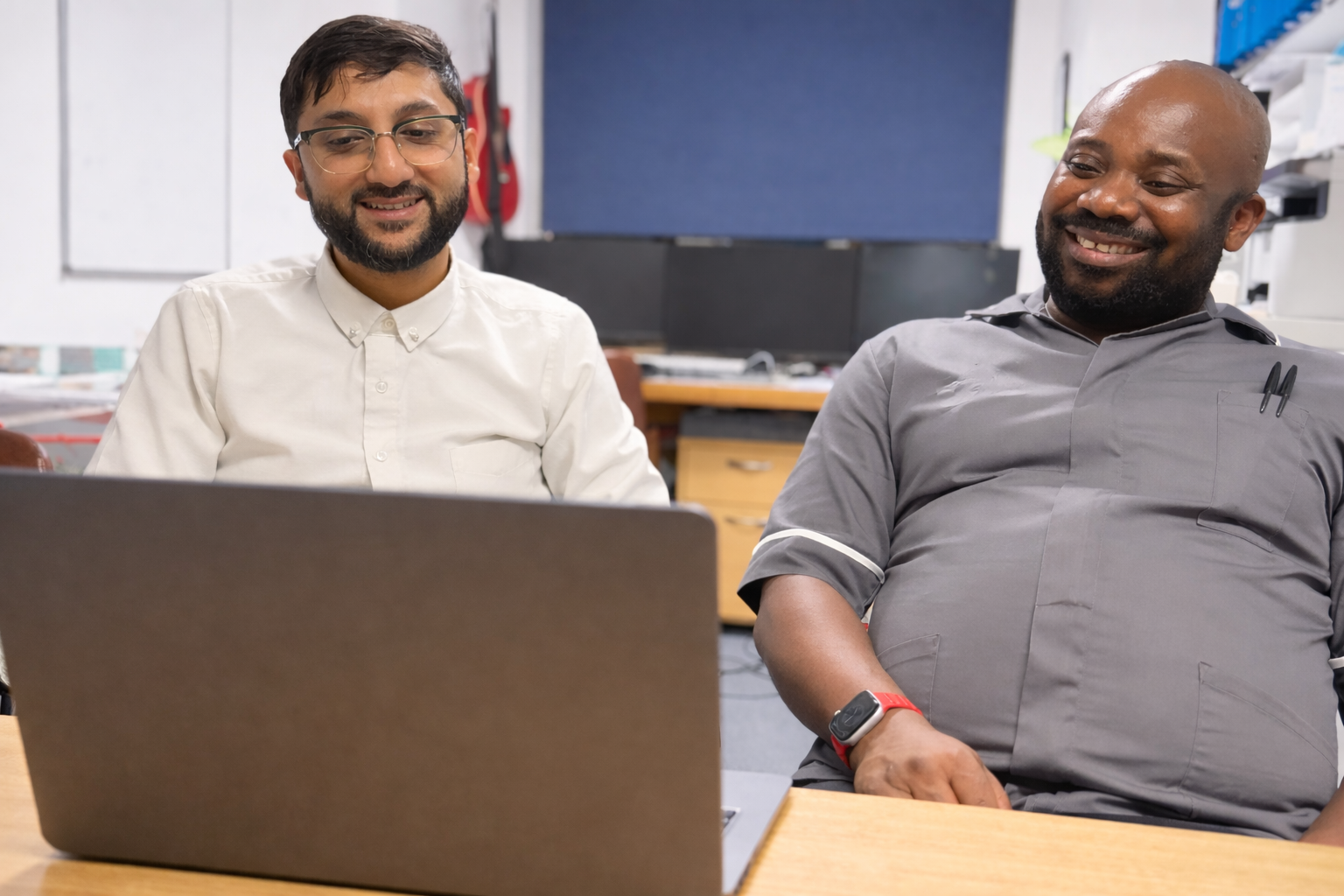 Two men sitting at a desk looking at a laptop, smiling indoors.