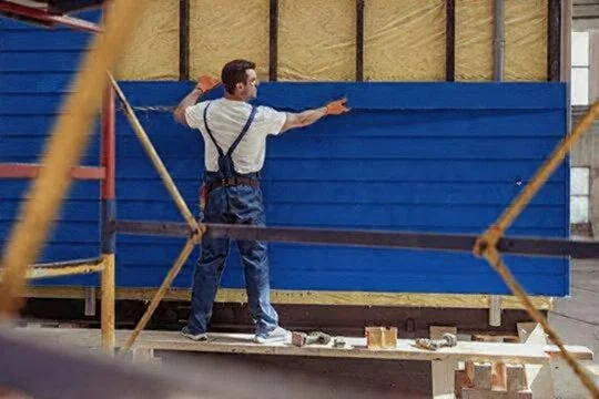 A man in work clothes painting a blue wooden wall inside a construction site.