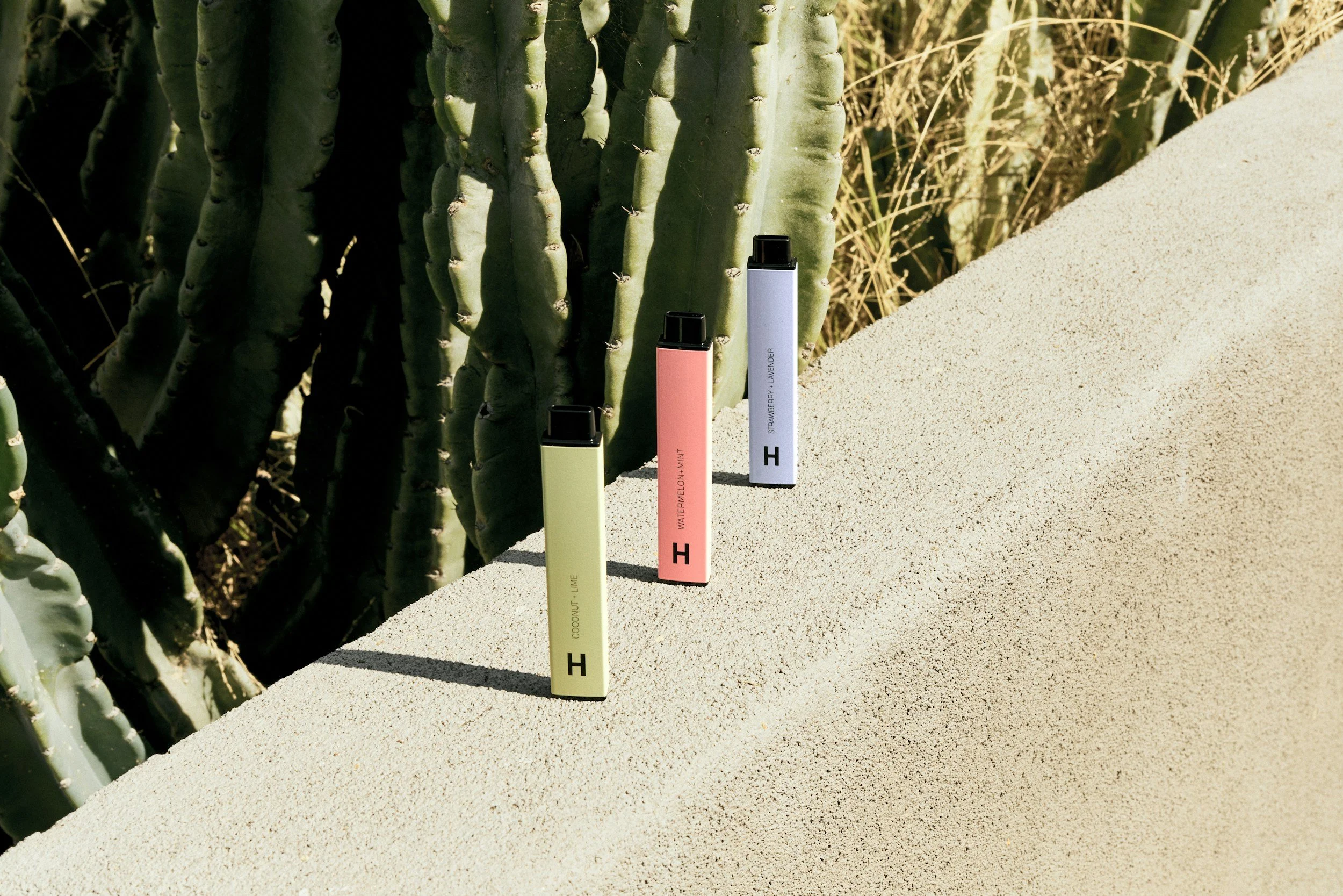 Three colorful vaporizer pens standing on a sandy surface with cactus plants in the background.