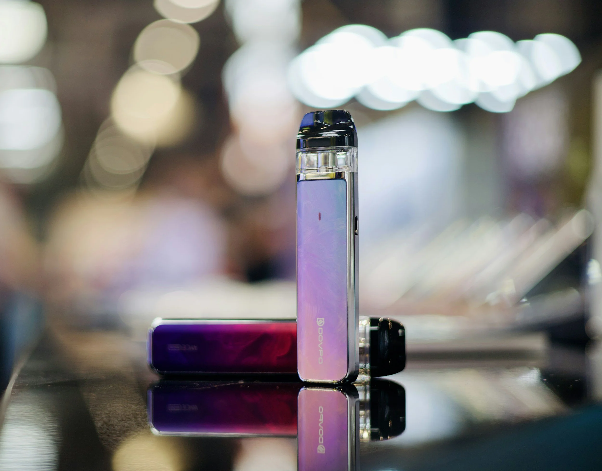 Two vape devices, one standing upright and the other lying down, on a reflective surface with a blurred background of lights.