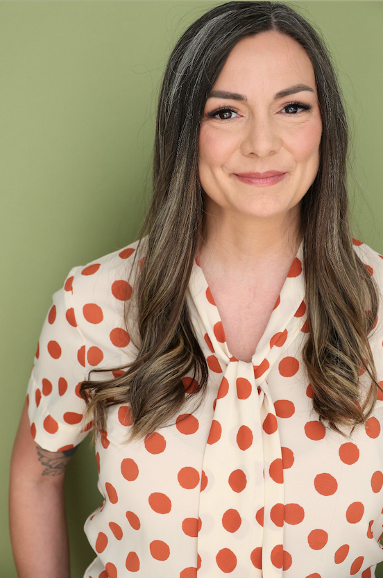 A woman with long brown hair and light makeup, smiling softly, wearing a cream blouse with red polka dots and a tied bow at the collar, standing against a green background.