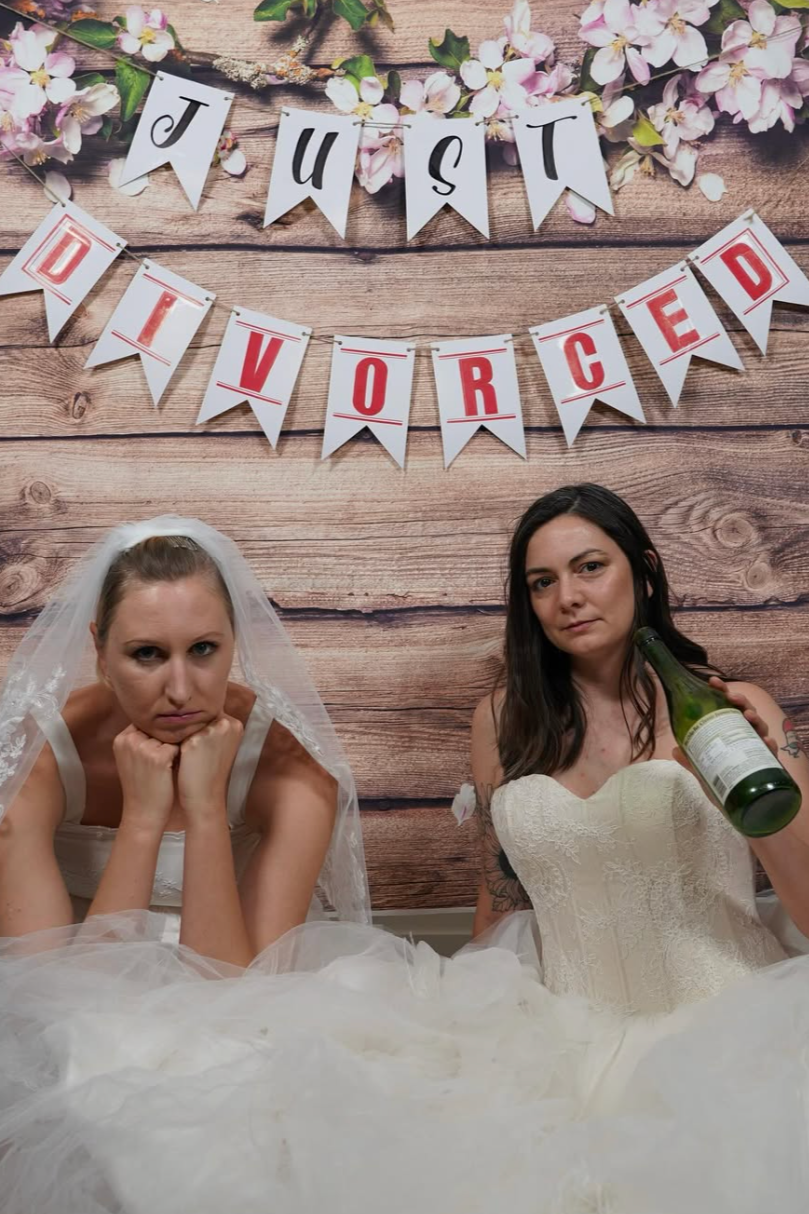 two women sit in wedding dresses, the woman on the left is disappointed with her head in her hands. The woman on the right looks snarky, a champagne bottle in her hand. Above them is a traditional "Just Married" banner, but reads "Just Divorced!"