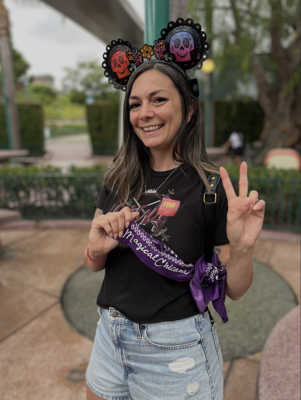 Woman smiling and making a peace sign while wearing a festive Dia de los Muertos-themed headband with colorful skulls.