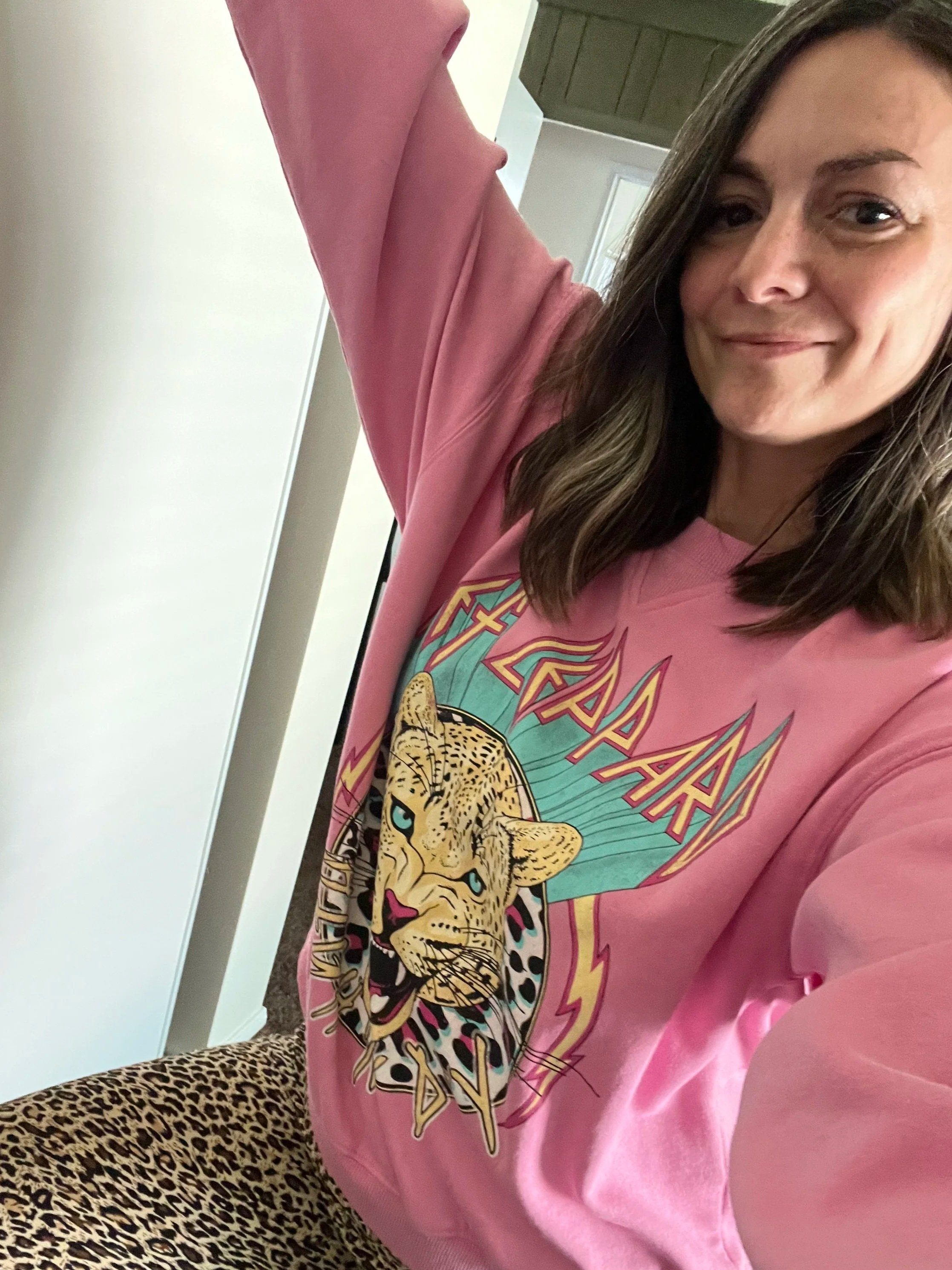 A woman with shoulder-length brown hair smiling and taking a selfie, wearing a pink graphic sweatshirt with a leaping jaguar and stylized text, and leopard print pants.