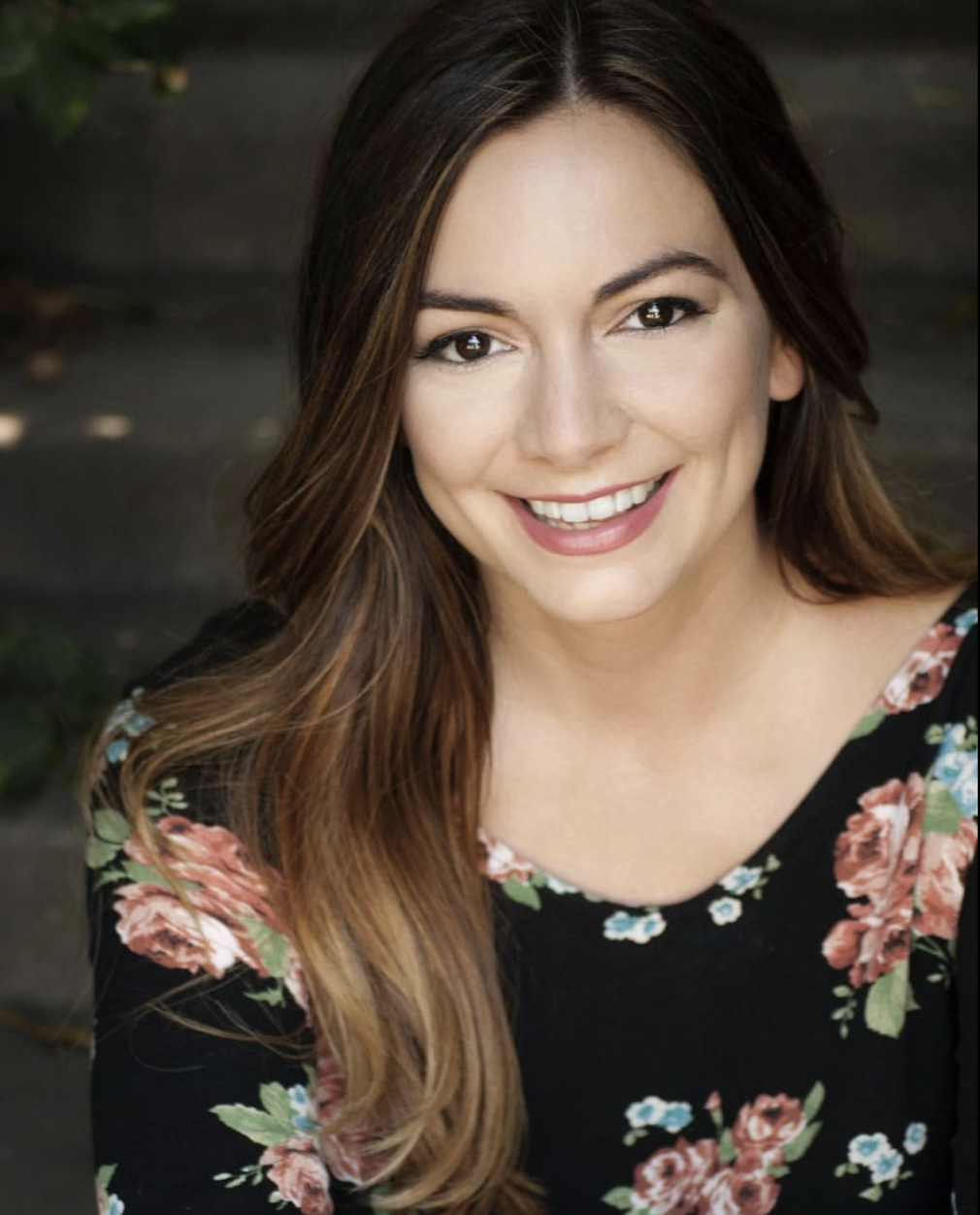 Close-up of a smiling woman with long, wavy brown hair, wearing a black floral top, outdoors with blurred dark background.