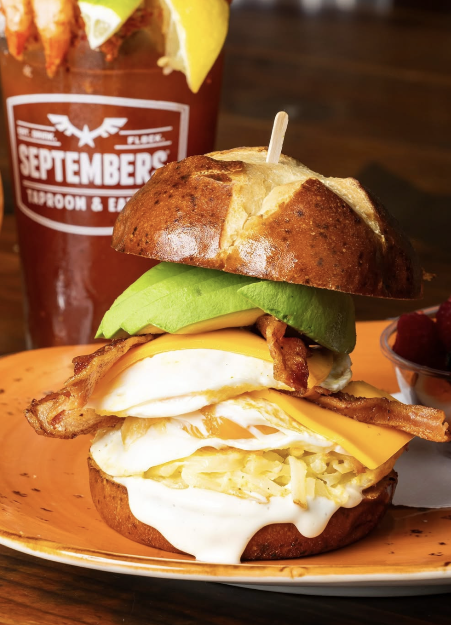 loaded bloody mary in the background, a stacked breakfast sandwich with egg, avocado, bacon and cheese on a pretzel bun