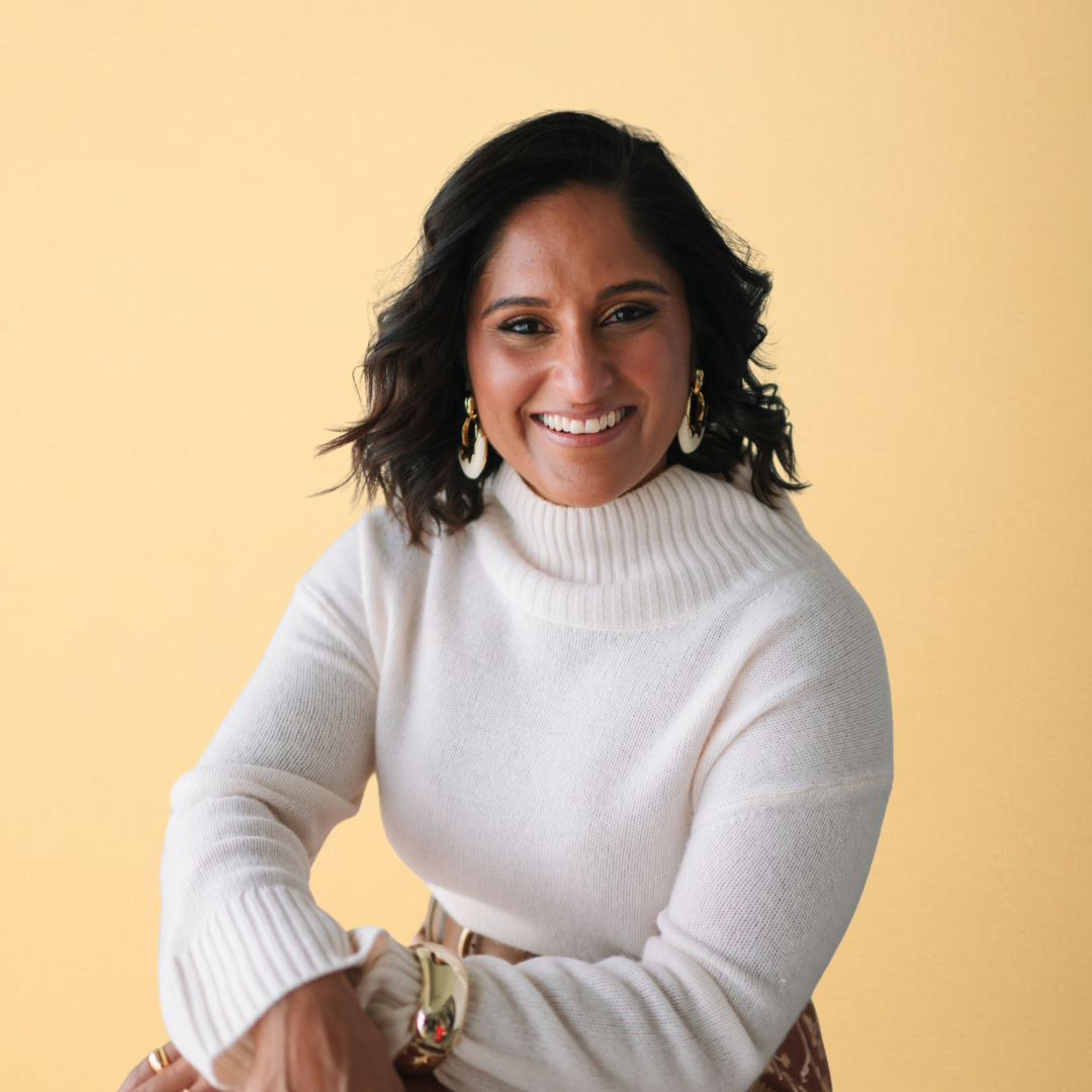 A woman with dark, wavy hair and medium skin tone smiling, wearing a white turtleneck sweater, large earrings, and a watch, with a yellow background.