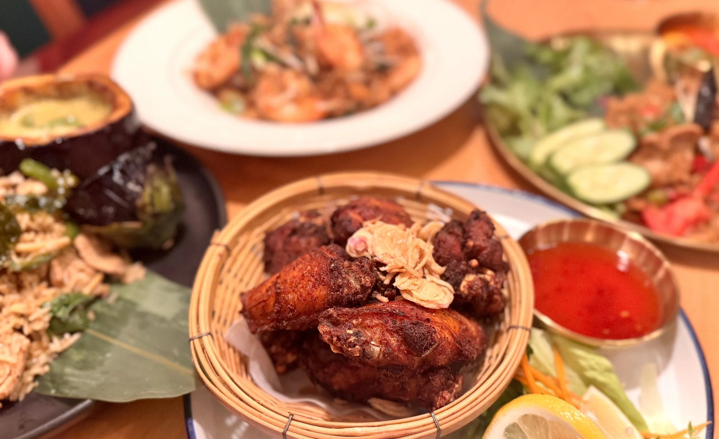 Assorted Middle Eastern or Mediterranean dishes including fried chicken wings, grilled vegetables, salads with cucumbers and tomatoes, rice dish, and a bowl of red dipping sauce on a wooden table.