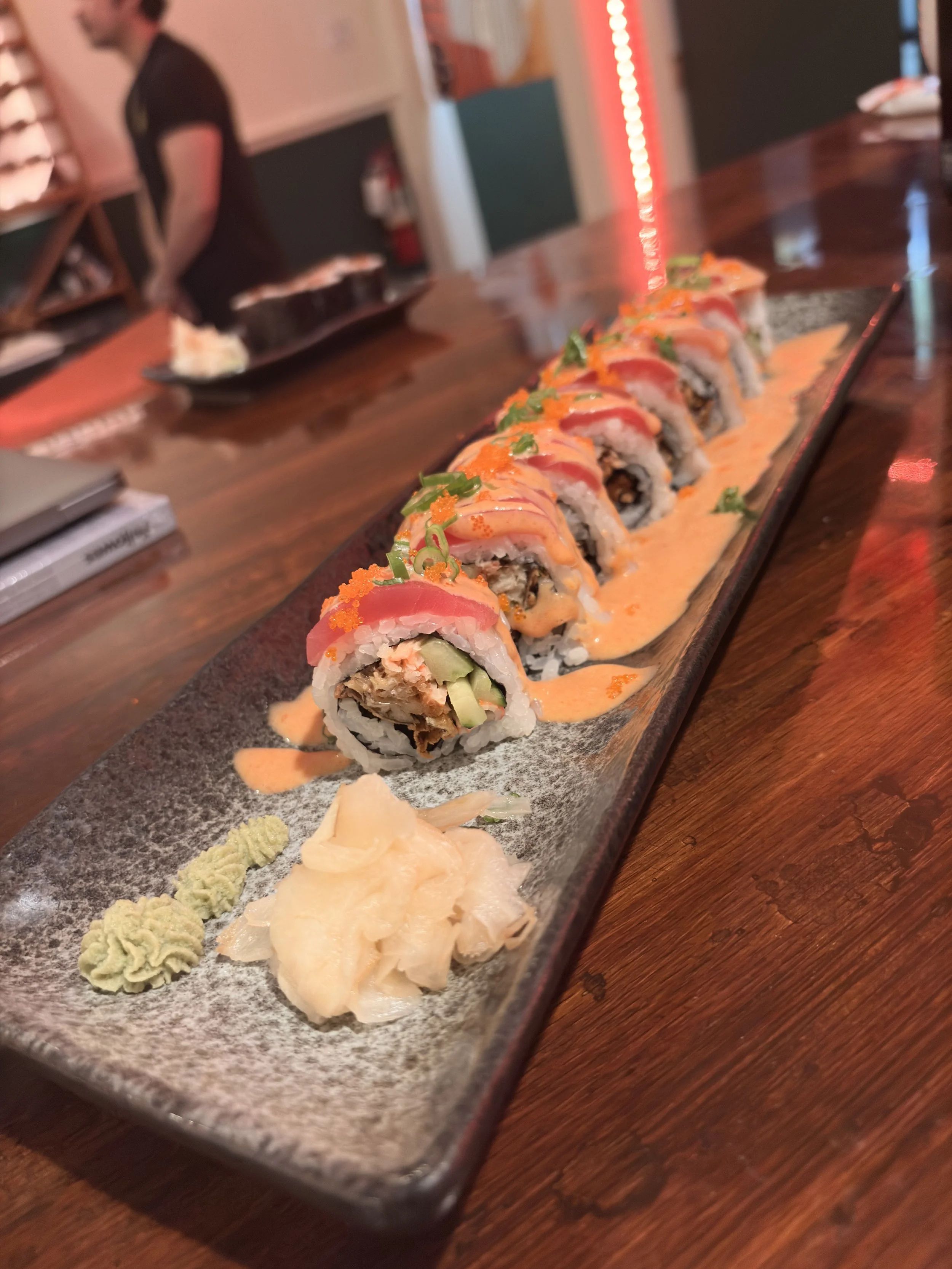 A long rectangular platter of sushi rolls topped with sliced fish, drizzled with sauce and garnished with green onions, accompanied by wasabi and pickled ginger.
