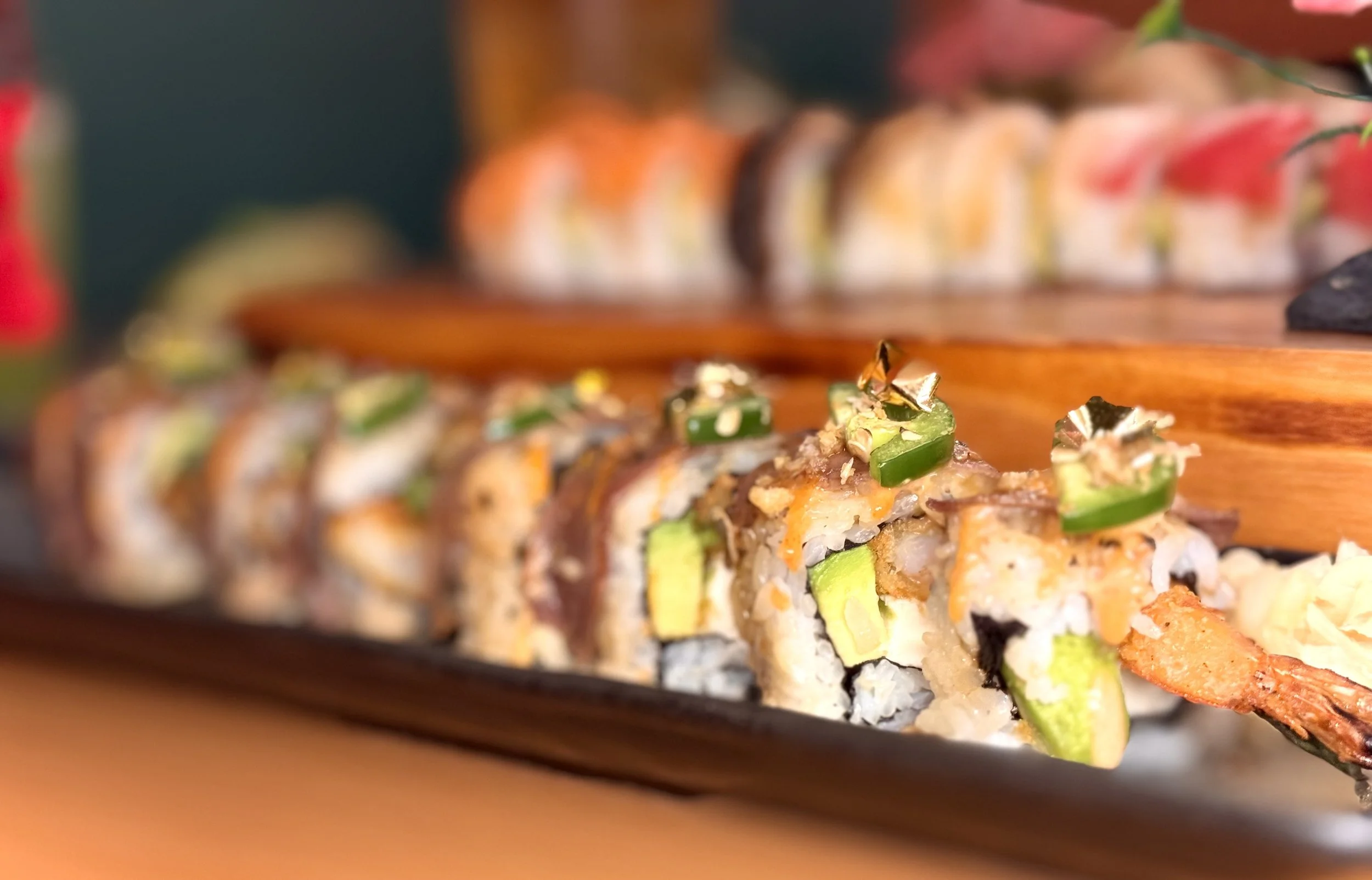 Close-up of assorted sushi rolls with various ingredients, garnished with green chili slices and gold leaf, on a black serving platter with a blurred background.