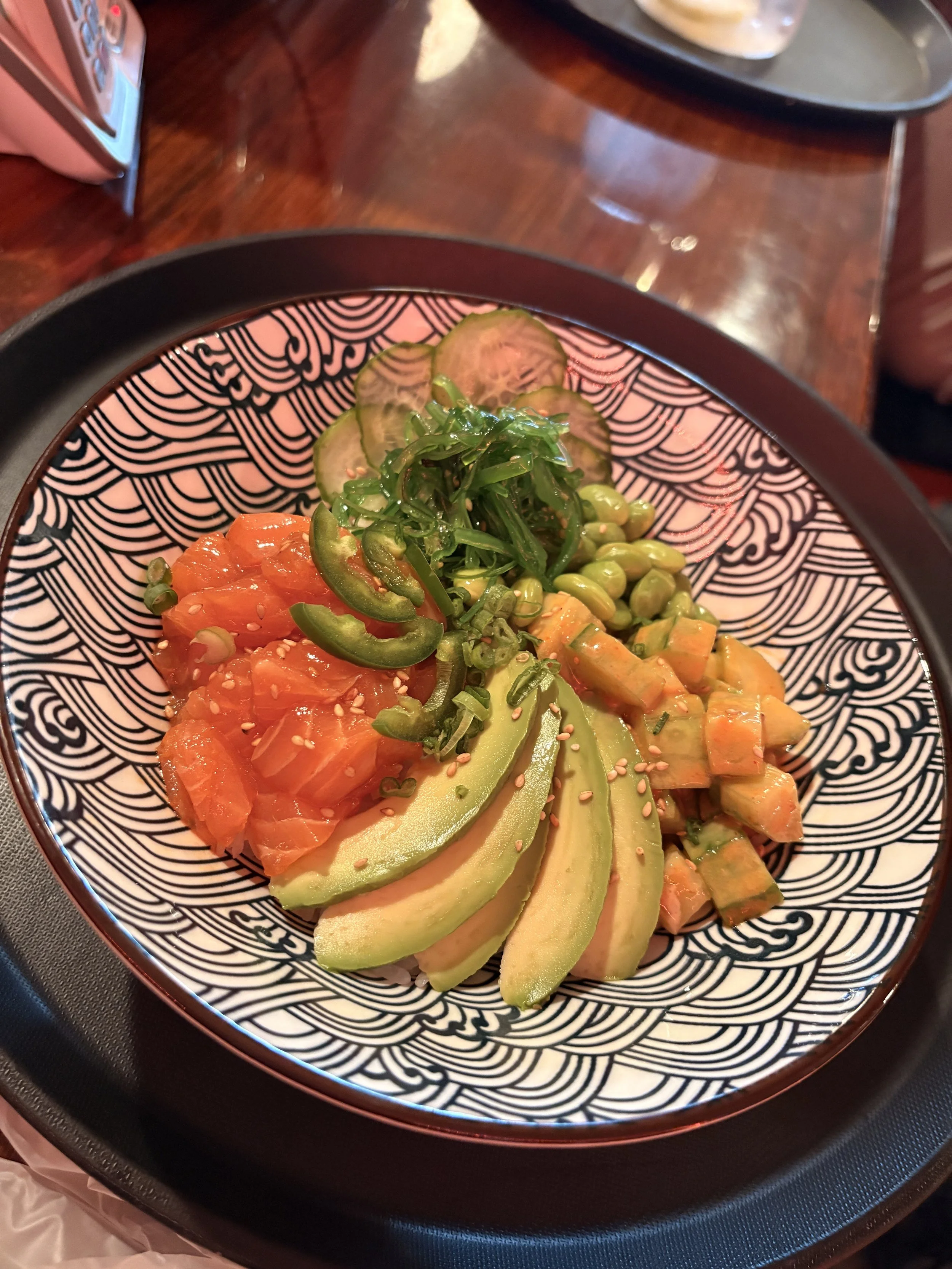 A bowl of assorted fresh vegetables including avocado slices, chopped tomatoes, cucumbers, green peppers, edamame, and seaweed, garnished with sesame seeds, on a patterned black and white dish.