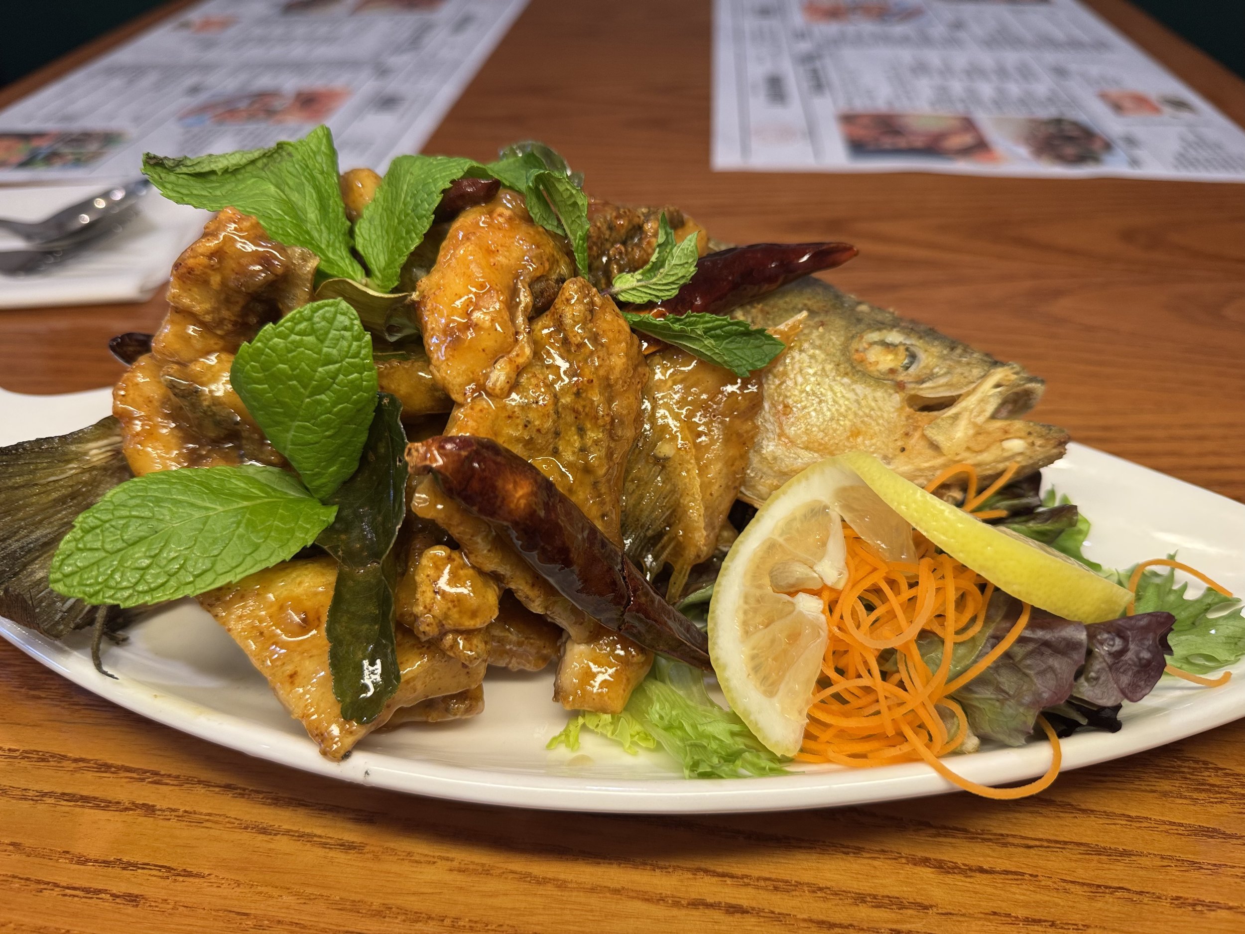A plate of fried fish garnished with green mint leaves and red dried chili peppers, served with a side salad including lemon wedges, shredded carrots, and mixed greens.