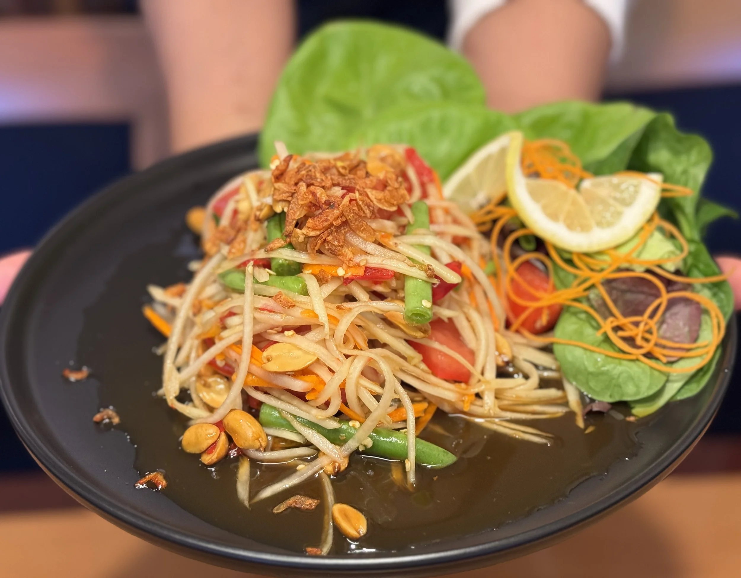 A black plate holding a serving of Thai papaya salad, garnished with crushed peanuts and fried garlic, accompanied by fresh lettuce, lemon wedges, and thinly shredded carrots.