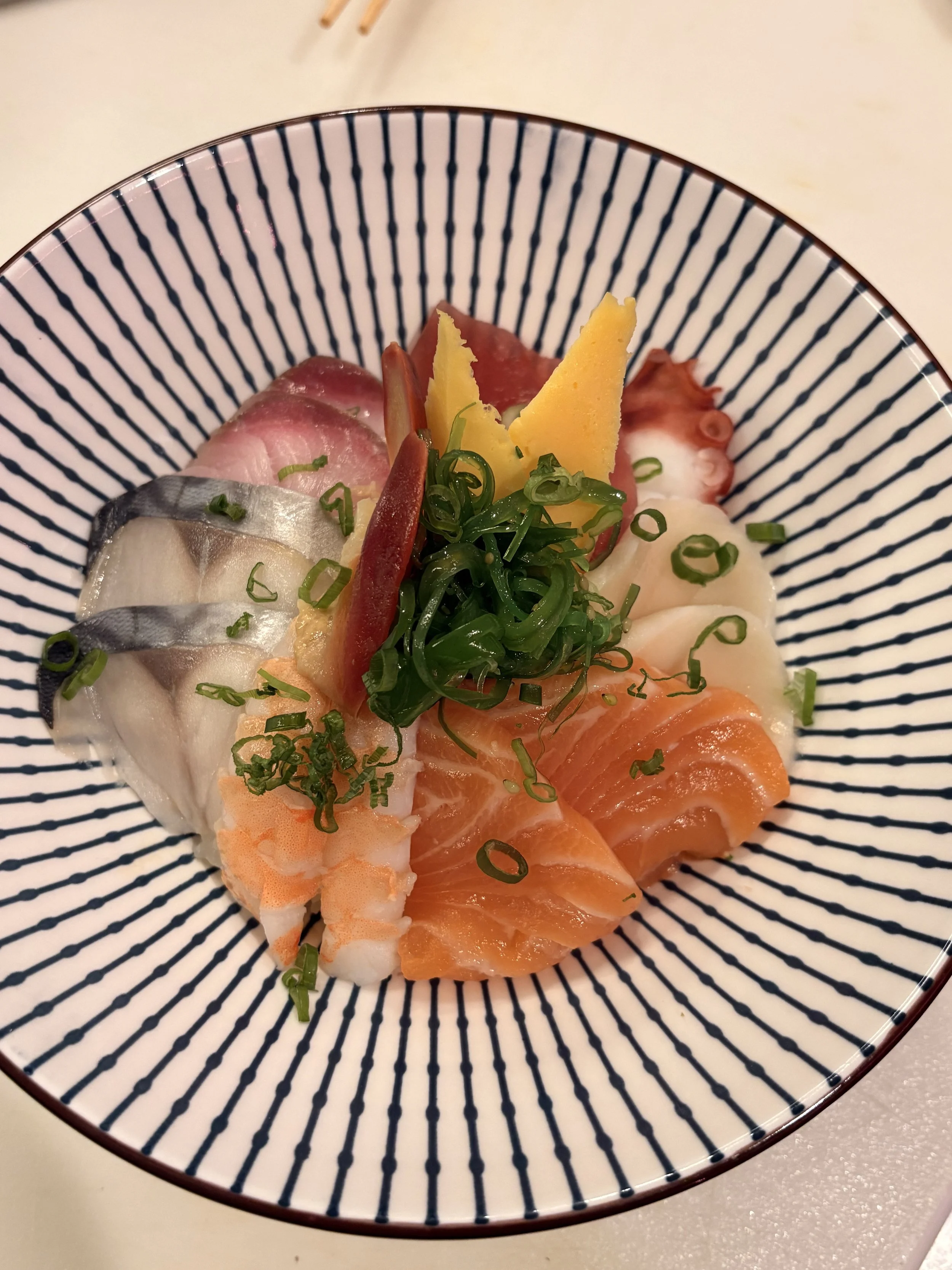 A bowl of assorted sashimi including slices of salmon, tuna, mackerel, shrimp, and octopus, garnished with green onions and seaweed.