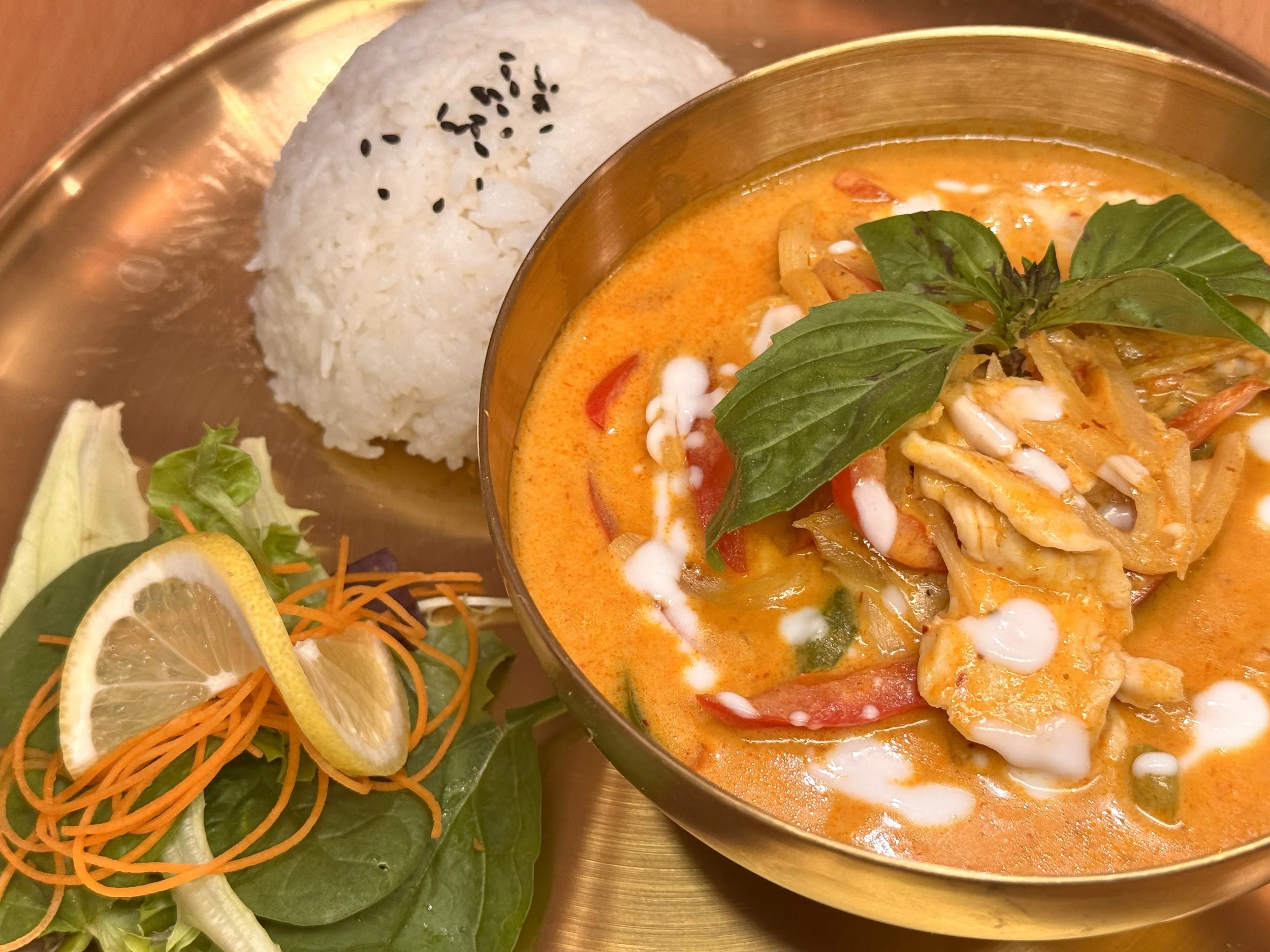 A bowl of Thai red curry with chicken, basil, and vegetables, served with white rice topped with black sesame seeds, and a side salad with lemon slice, lettuce, shredded carrots, and cucumber.