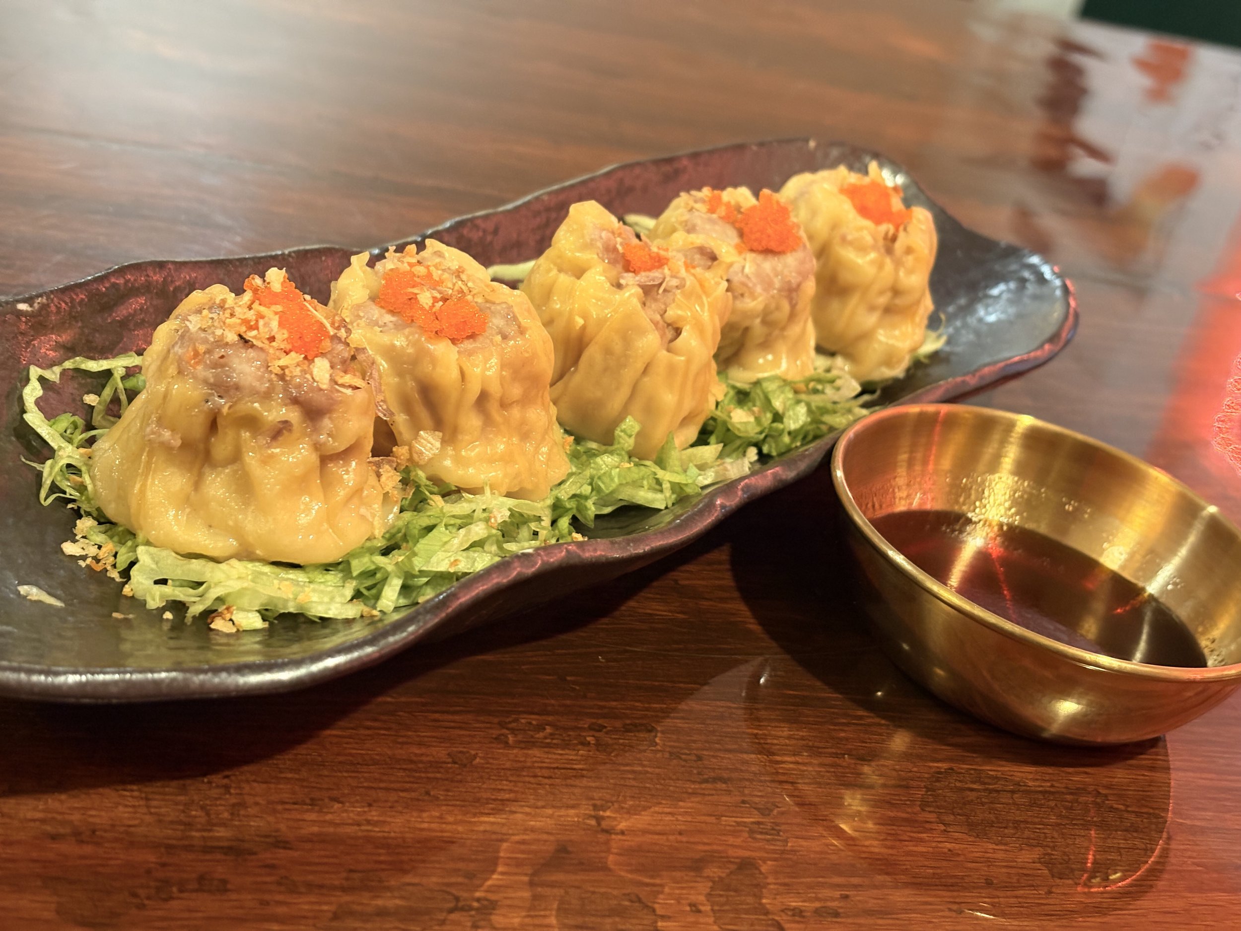 Five steamed dumplings on a black plate garnished with shredded lettuce and orange fish roe, with a small metallic bowl of dipping sauce on a wooden table.