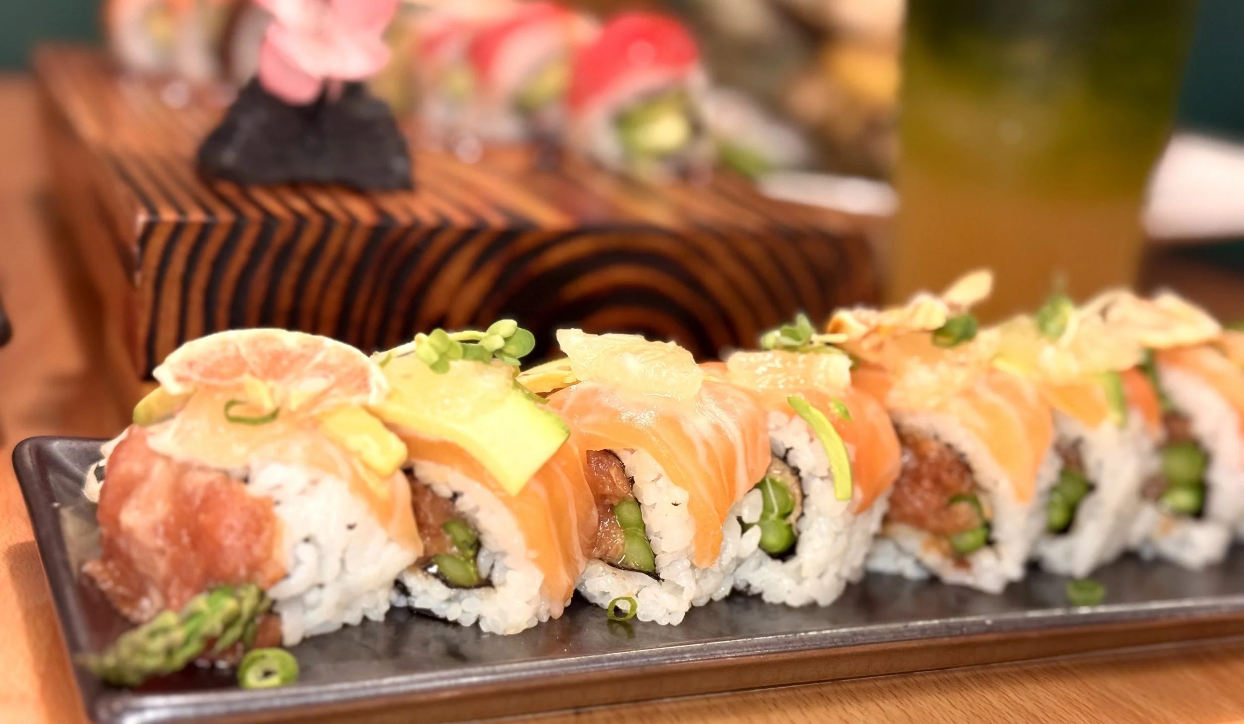 Close-up of a sushi roll topped with salmon, avocado, and garnished with sprouts and citrus slices, served on a black rectangular plate.