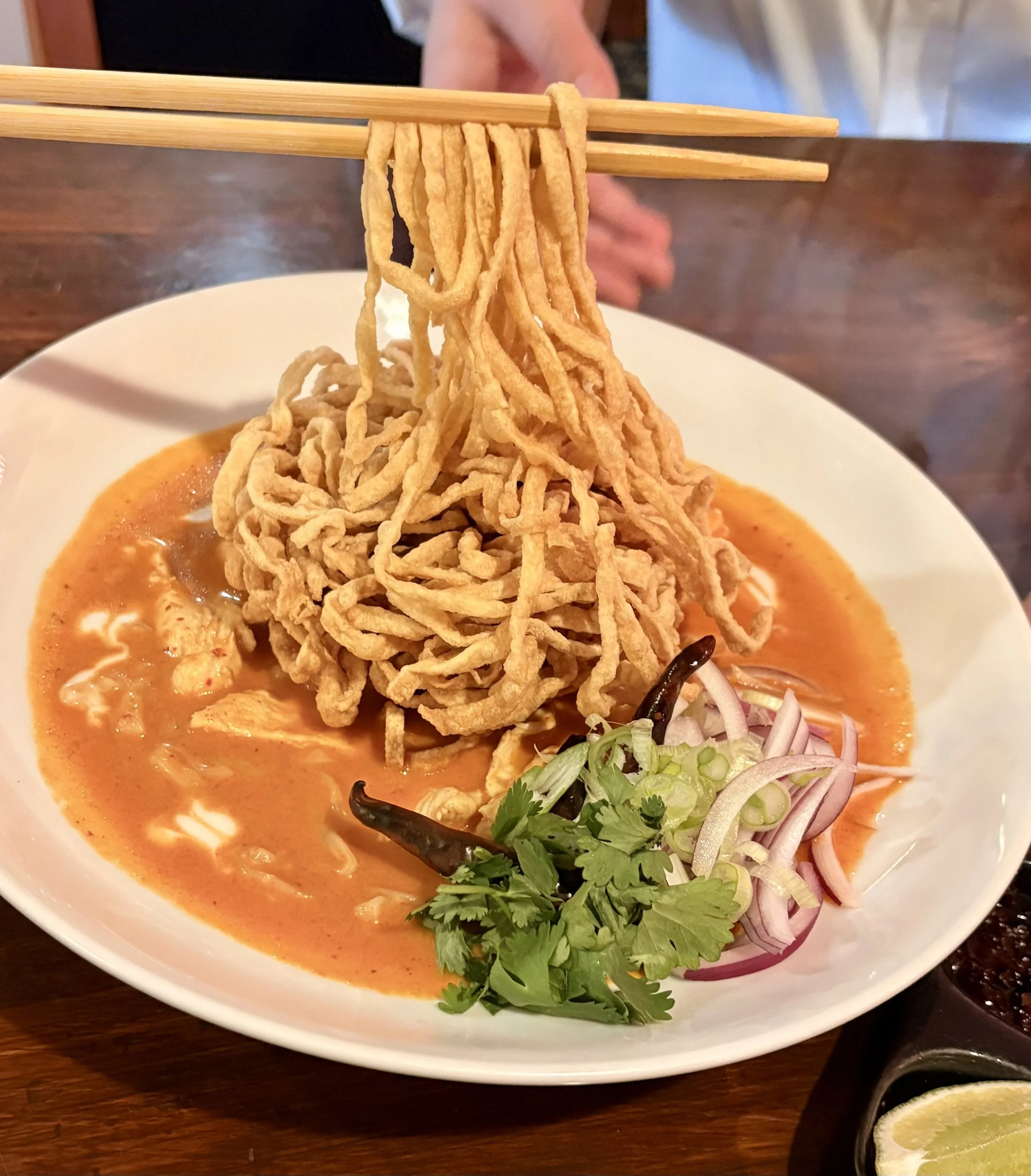 A bowl of spicy red curry with fried noodles, garnished with cilantro, red onion, and chili peppers, served on a wooden table.
