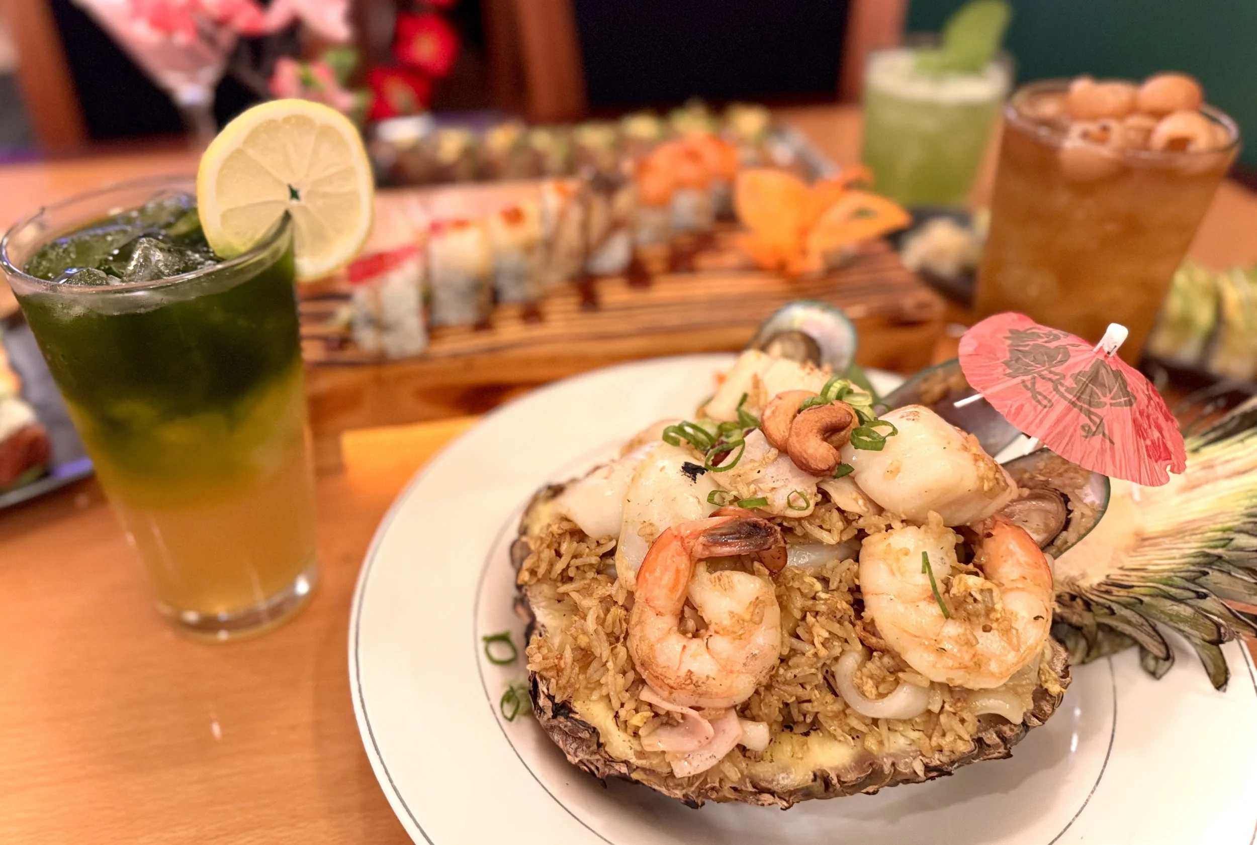 A plate of seafood fried rice served inside a hollowed pineapple, garnished with cashews and green onions, with a small pink paper umbrella on top. In the background, there are two drinks with ice, one with a lemon slice and green color, and the othe