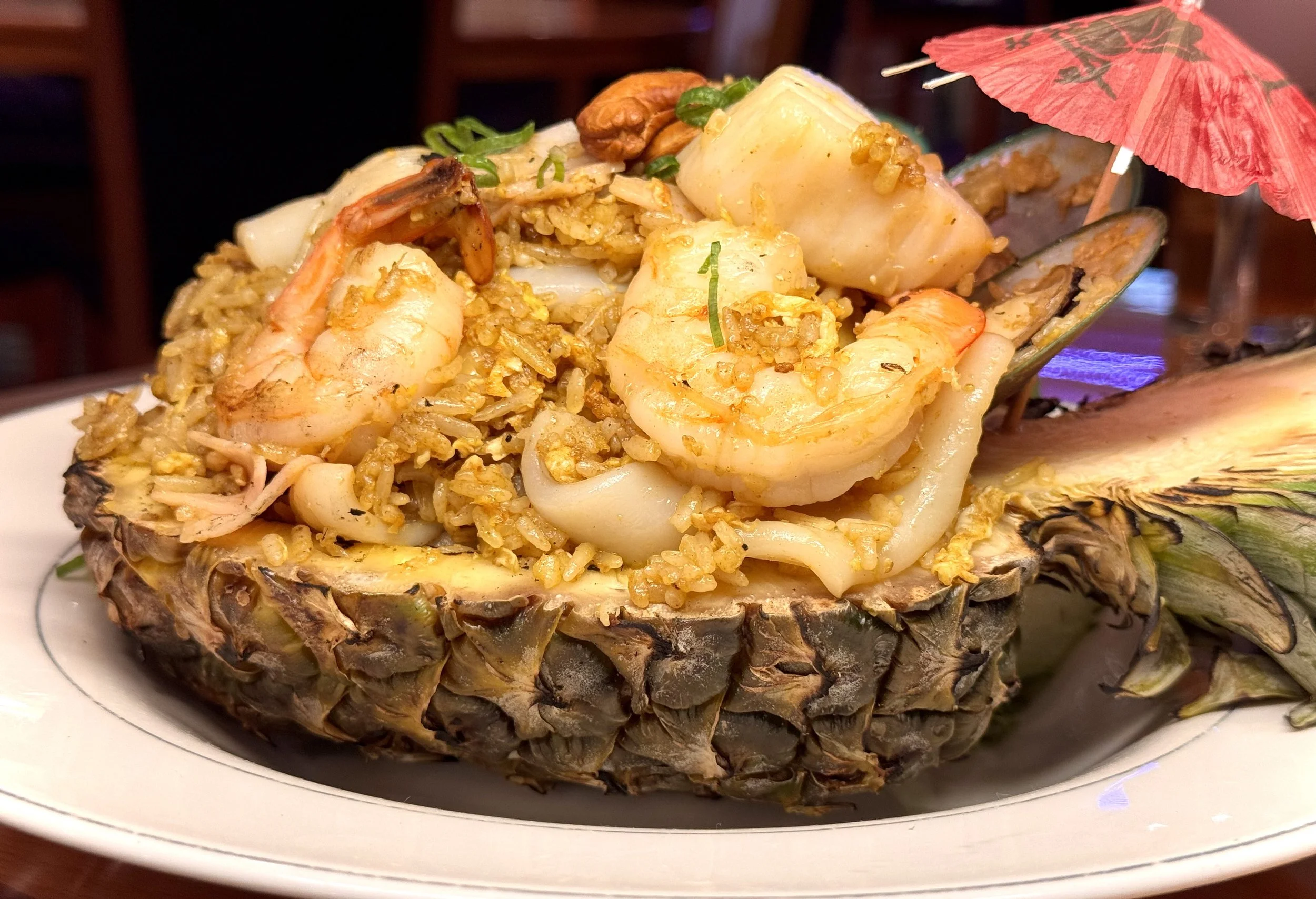 A pineapple half filled with fried rice, shrimp, squid, and garnished with a small umbrella and green onions.