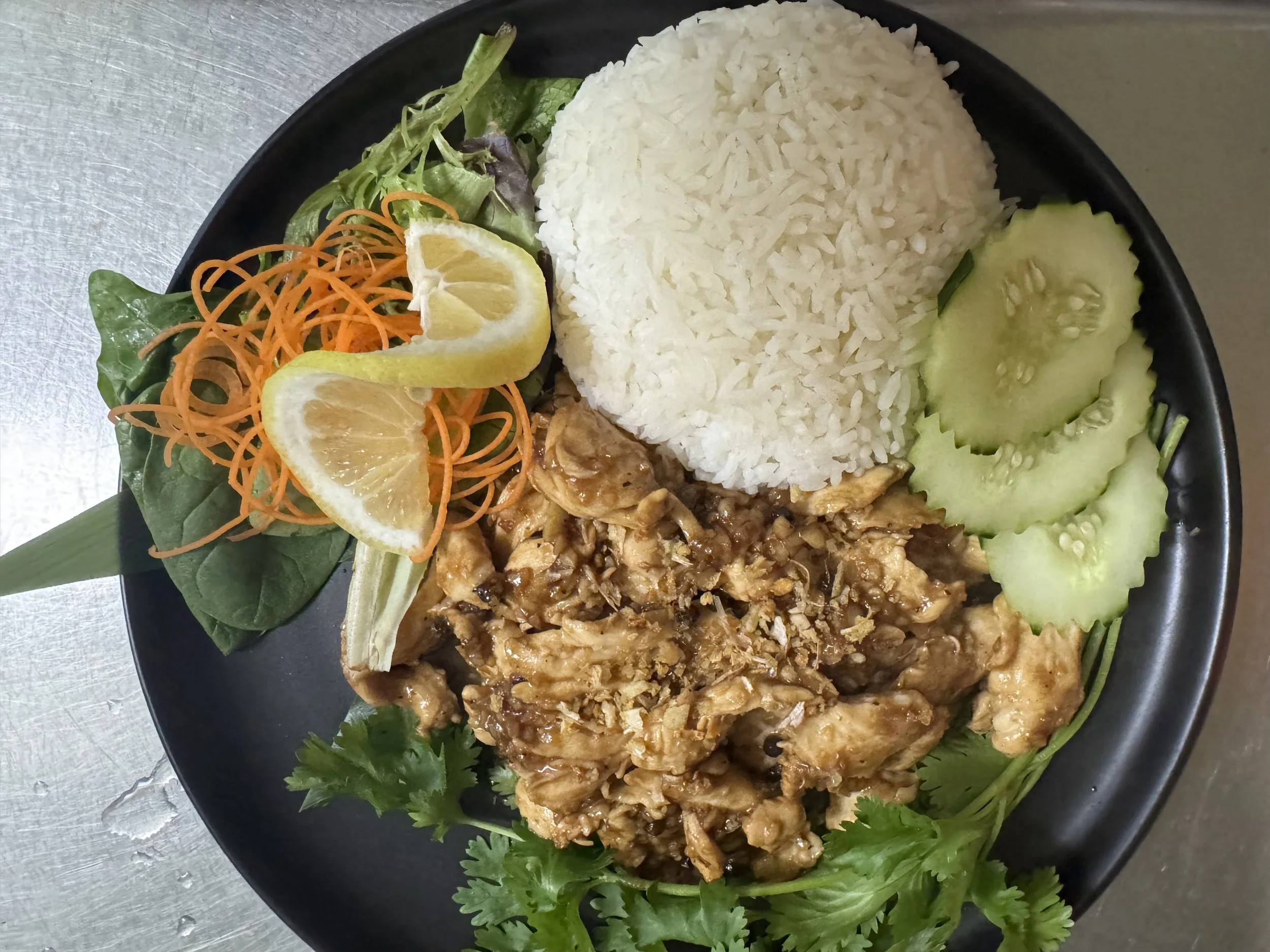 Plate of food with white rice, cucumber slices, cooked chicken with sauce, fresh cilantro, lemon slices, shredded carrots, and leafy greens.