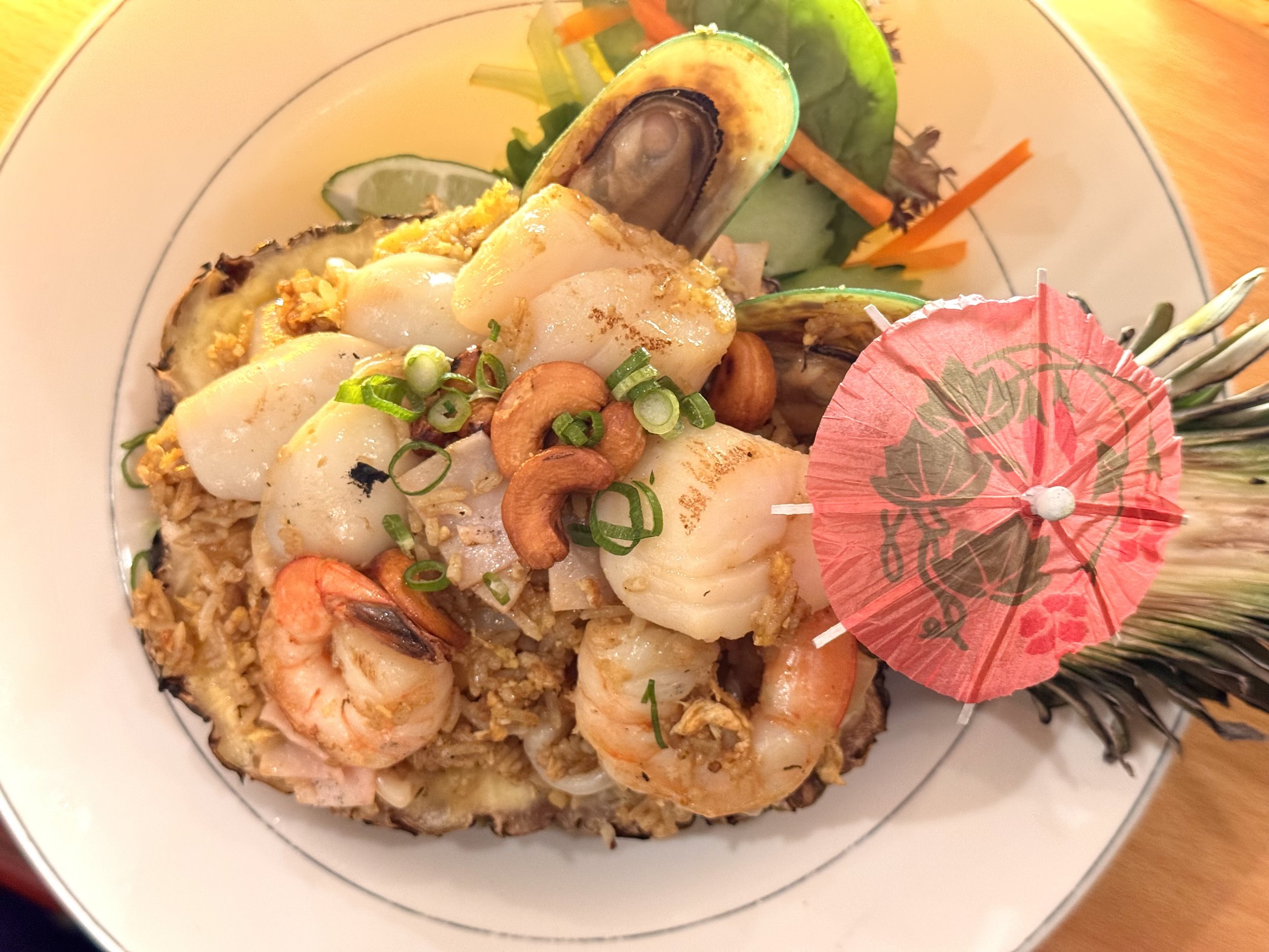 Seafood dish with shrimp, squid, mussels, and rice, garnished with cashews, chopped green onions, and a small pink paper umbrella, served in a pineapple boat with vegetables and lime slice.