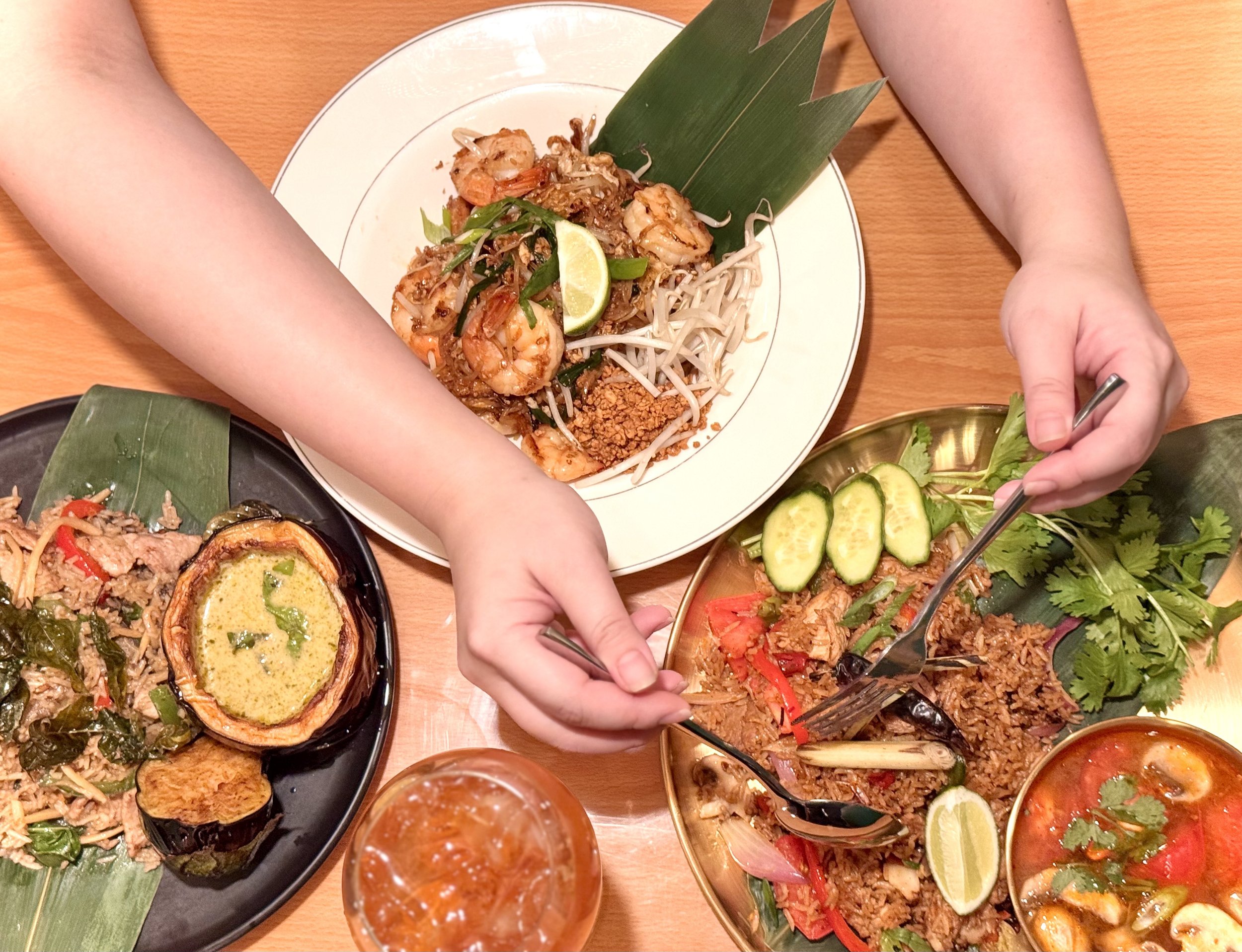 Three plates of Thai food on a wooden table with a drink, including a pad thai with shrimp, a spicy beef stir-fry, and a green curry with eggplant, cucumber, cilantro, and lime.