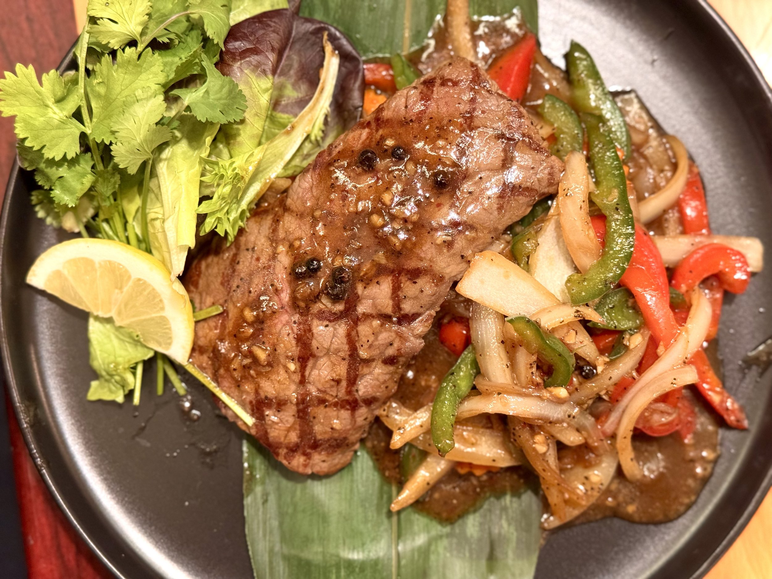Grilled steak with peppercorn sauce, served with sautéed bell peppers and onions, garnished with fresh cilantro and lemon wedges.