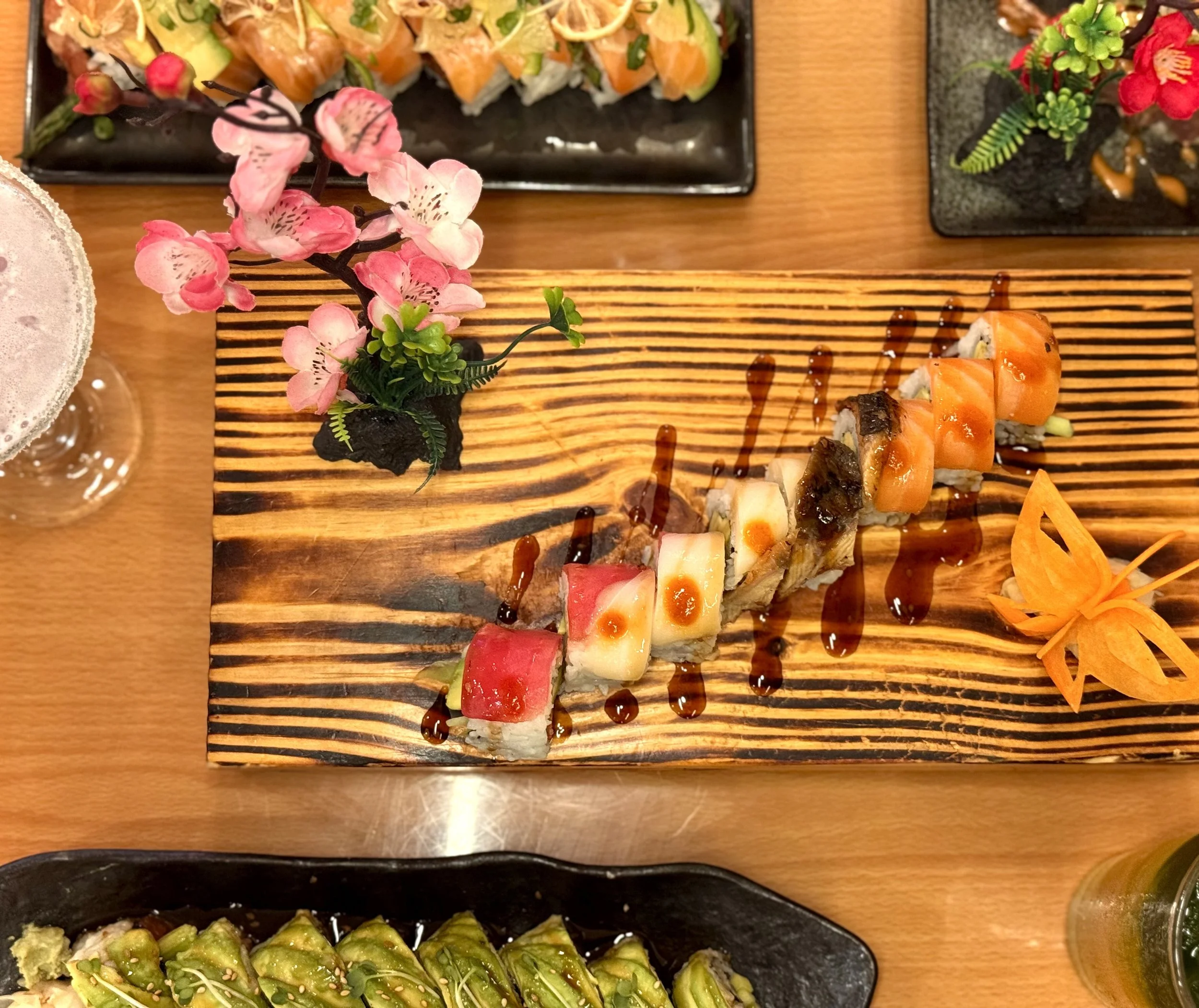 A wooden platter with assorted sushi rolls drizzled with sauce and decorated with a pink and white artificial cherry blossom branch. Two other plates with sushi and a drink are partially visible on a wooden table.