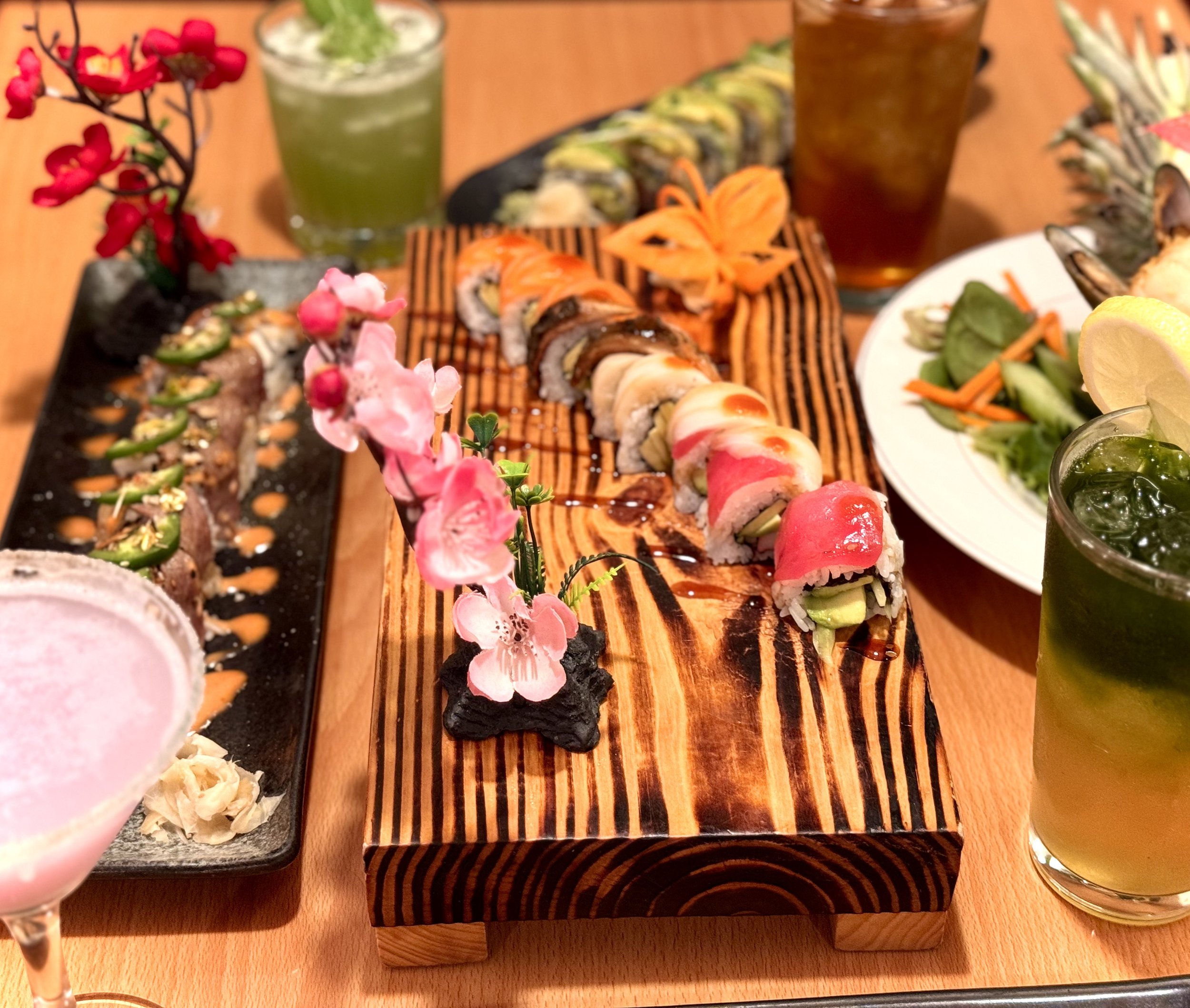 Assorted sushi rolls with various toppings on a wooden platter, surrounded by drinks and side dishes, including a salad and garnishes.