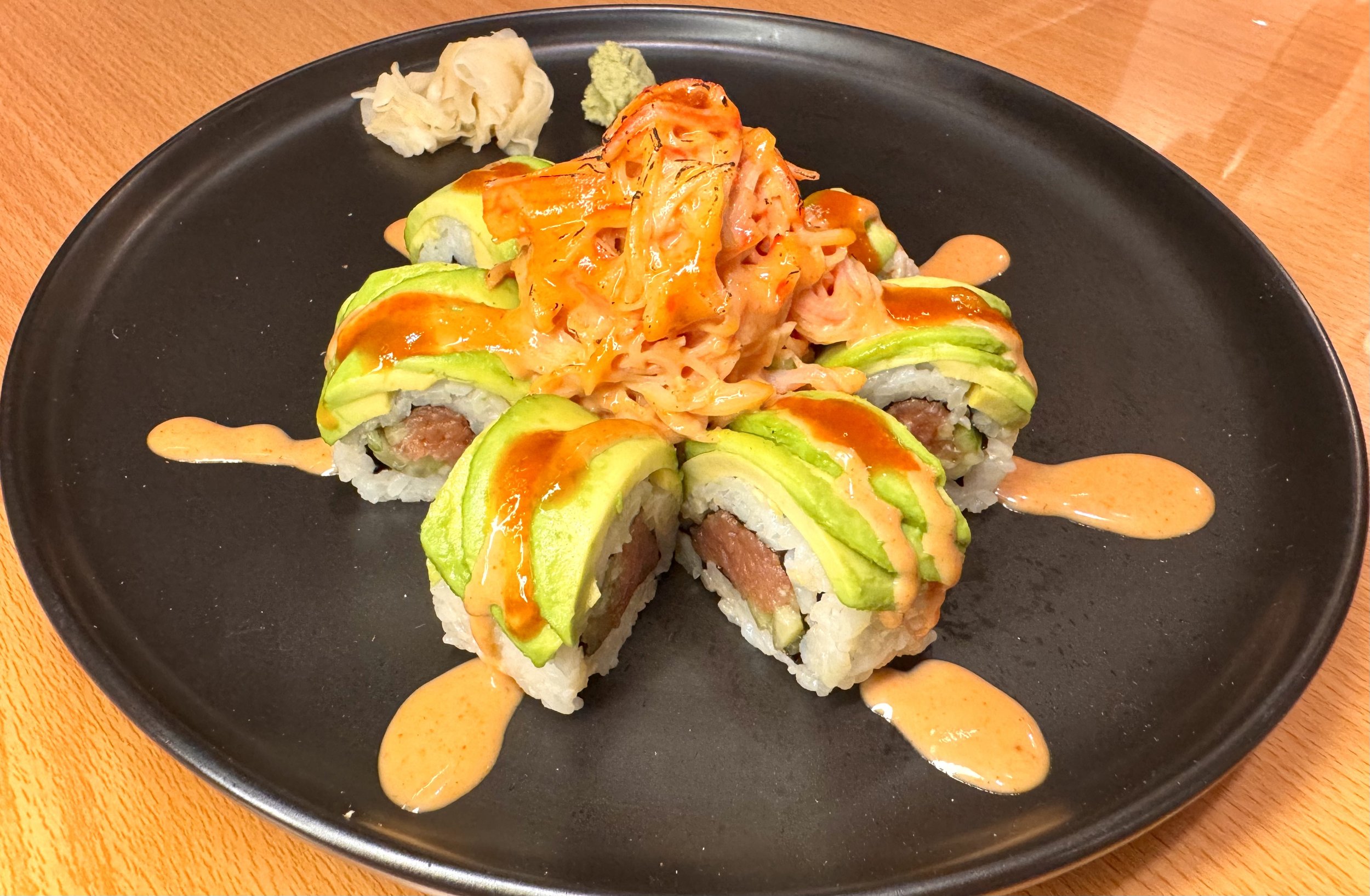 Sushi roll topped with avocado slices and drizzled with spicy orange sauce, with a small side of pickled ginger and wasabi on a black plate.