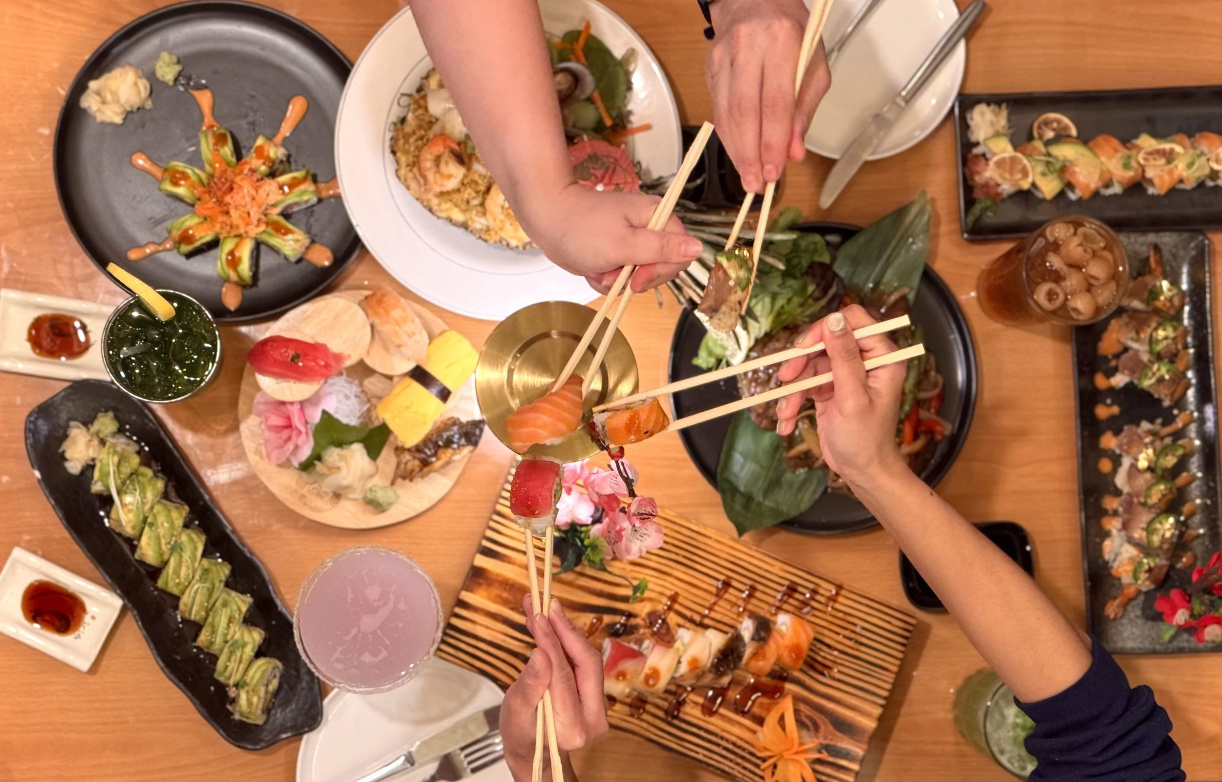A top-down view of a dining table filled with various Japanese dishes, sushi, drinks, and people using chopsticks to share food.