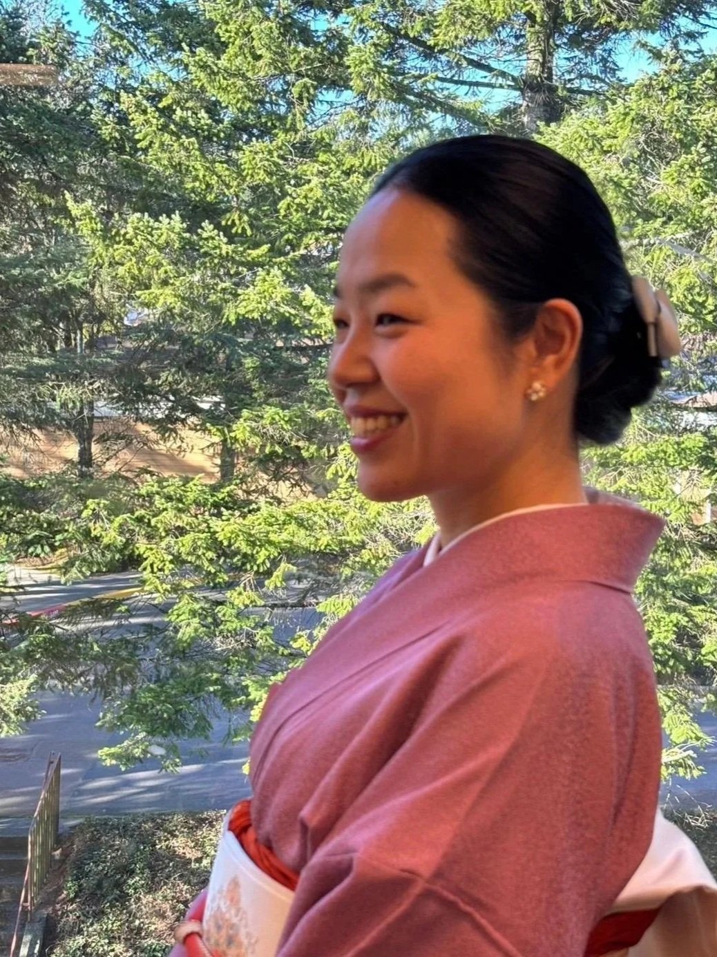 A woman dressed in traditional Japanese kimono with a smile, standing outdoors with green trees and a blue sky in the background.
