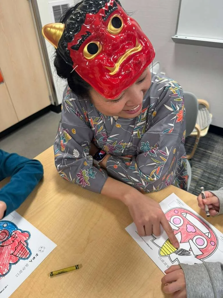 Person wearing a red demon mask with gold horns and black hair, sitting at a table and pointing to a coloring sheet of a similar masked face.